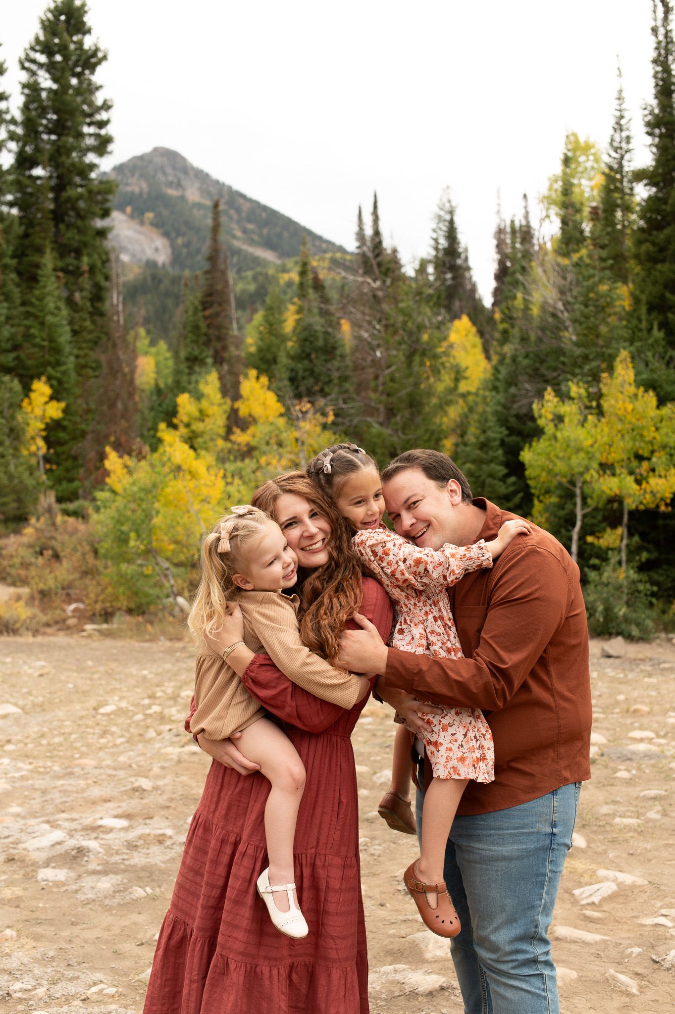 Family of four smiling outdoors in autumn mountain setting with colorful fall foliage and evergreen trees in background.