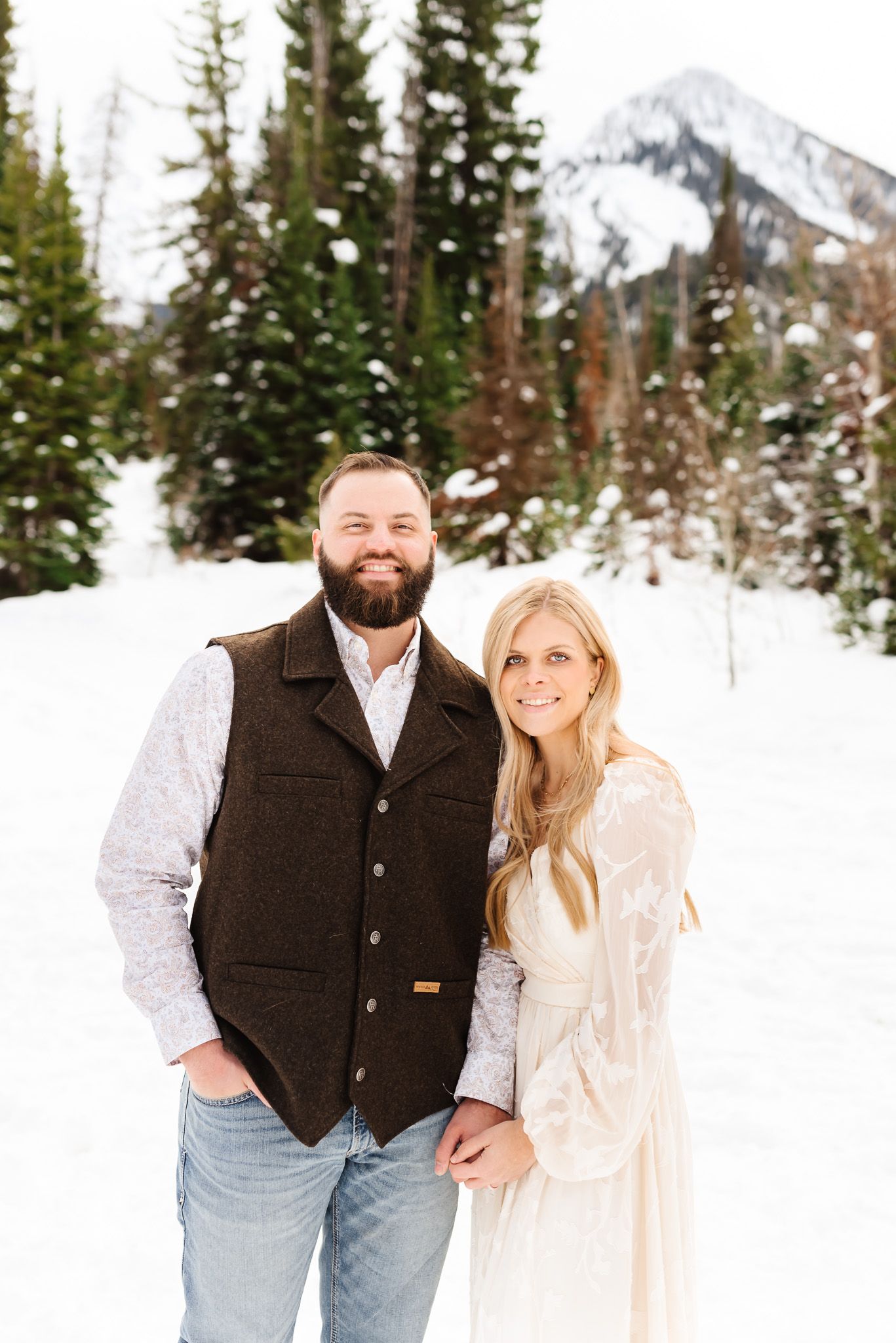 Happy couple posing together in casual winter attire against snowy mountain scenery.