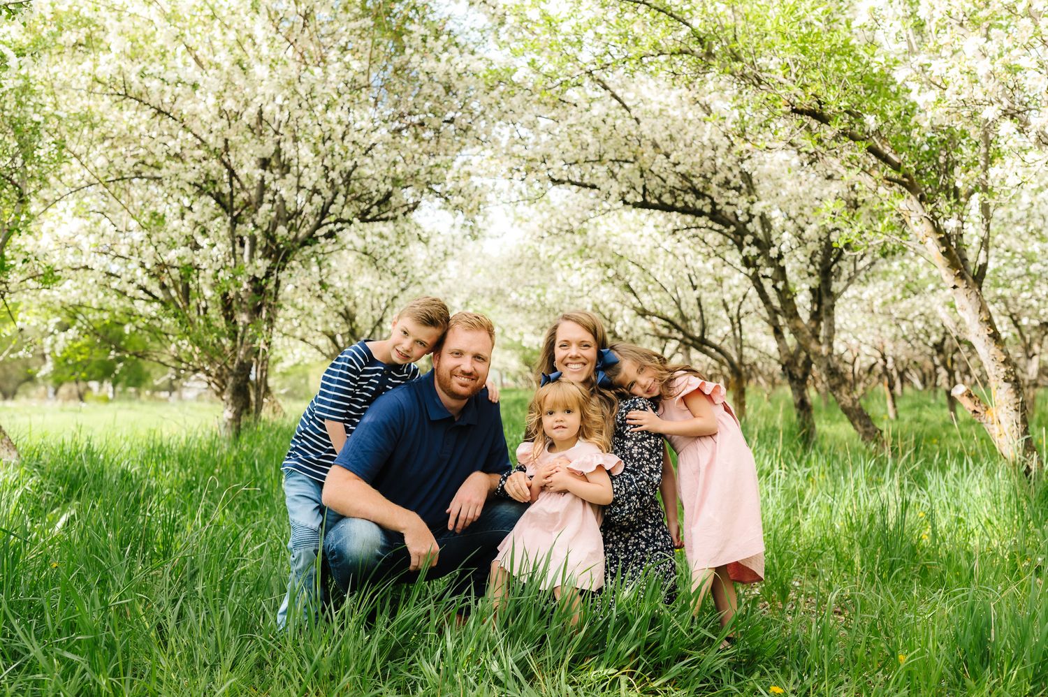 A joyful family photo session in a blooming spring orchard with white blossoms and lush green grass.
