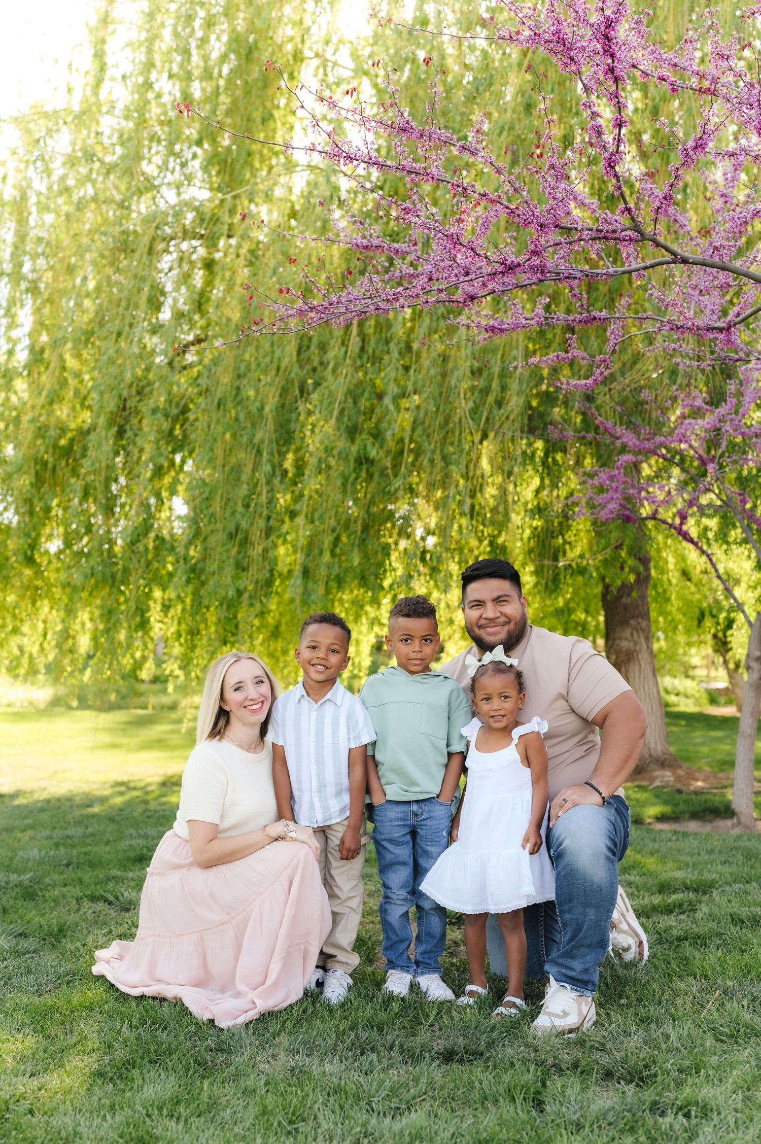 A joyful family sits together on grass under a blooming cherry blossom tree on a sunny spring day.