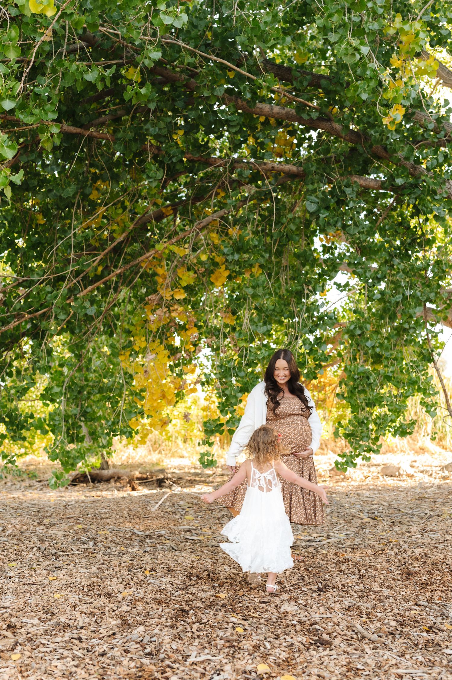 Pregnant woman in brown top smiles at young girl in white dress dancing under a large green tree outdoors.