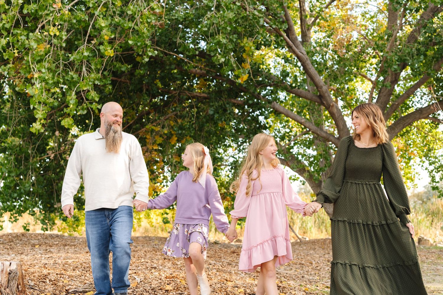 Family of four holding hands walking outdoors under green trees on a sunny day.