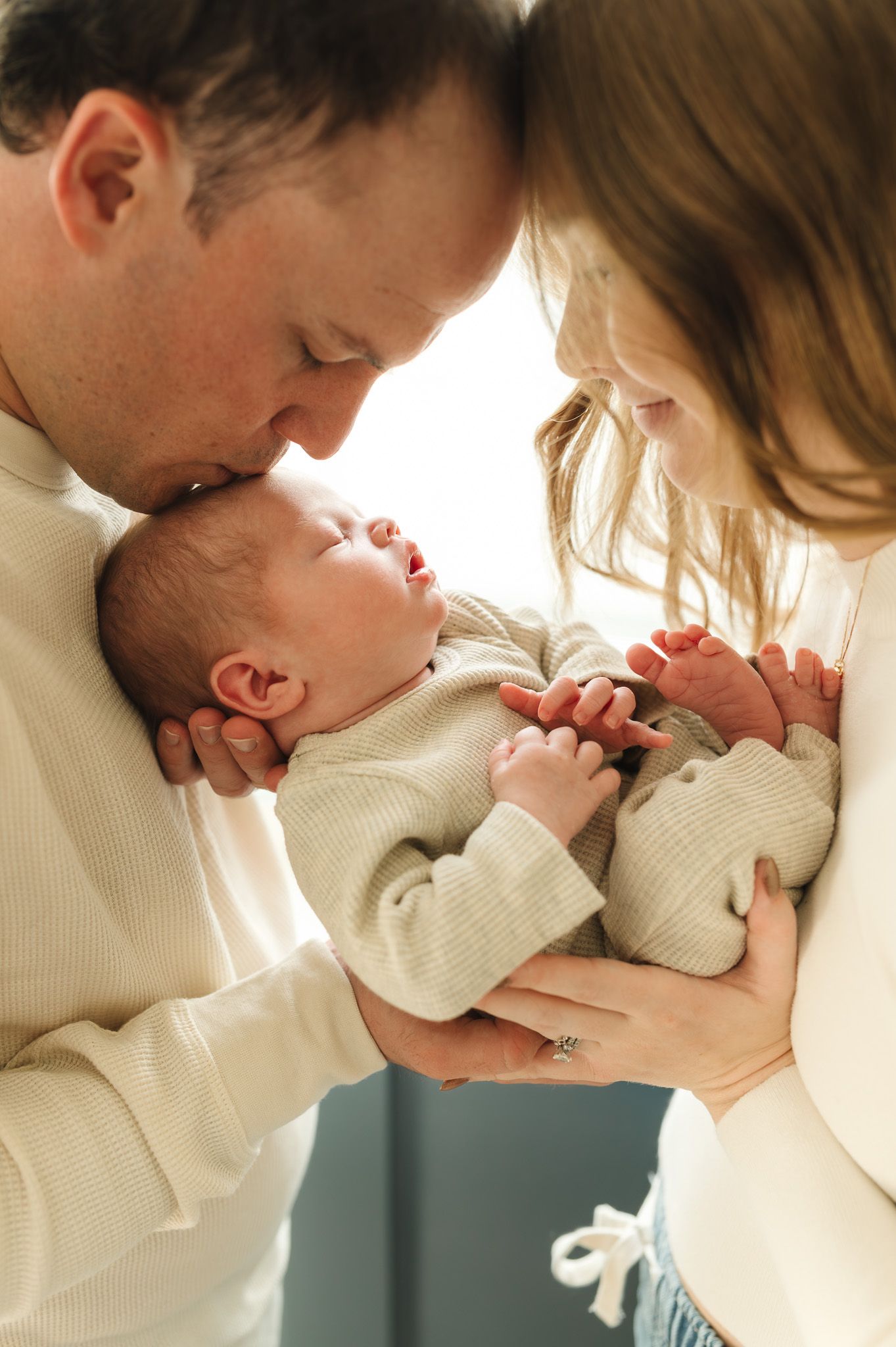 New parents tenderly kissing and cradling their sleeping newborn baby dressed in a cream outfit.
