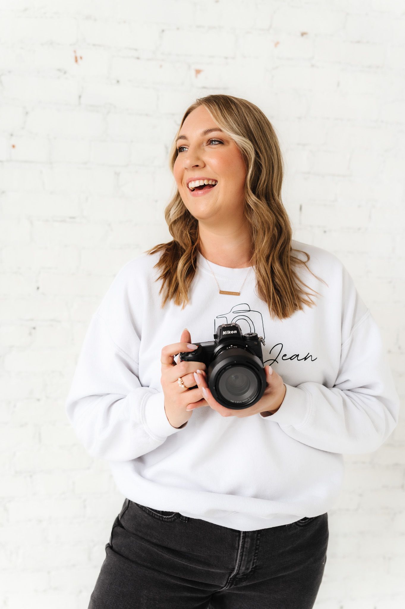 Photographer wearing white sweatshirt laughs while holding DSLR camera in studio setting.
