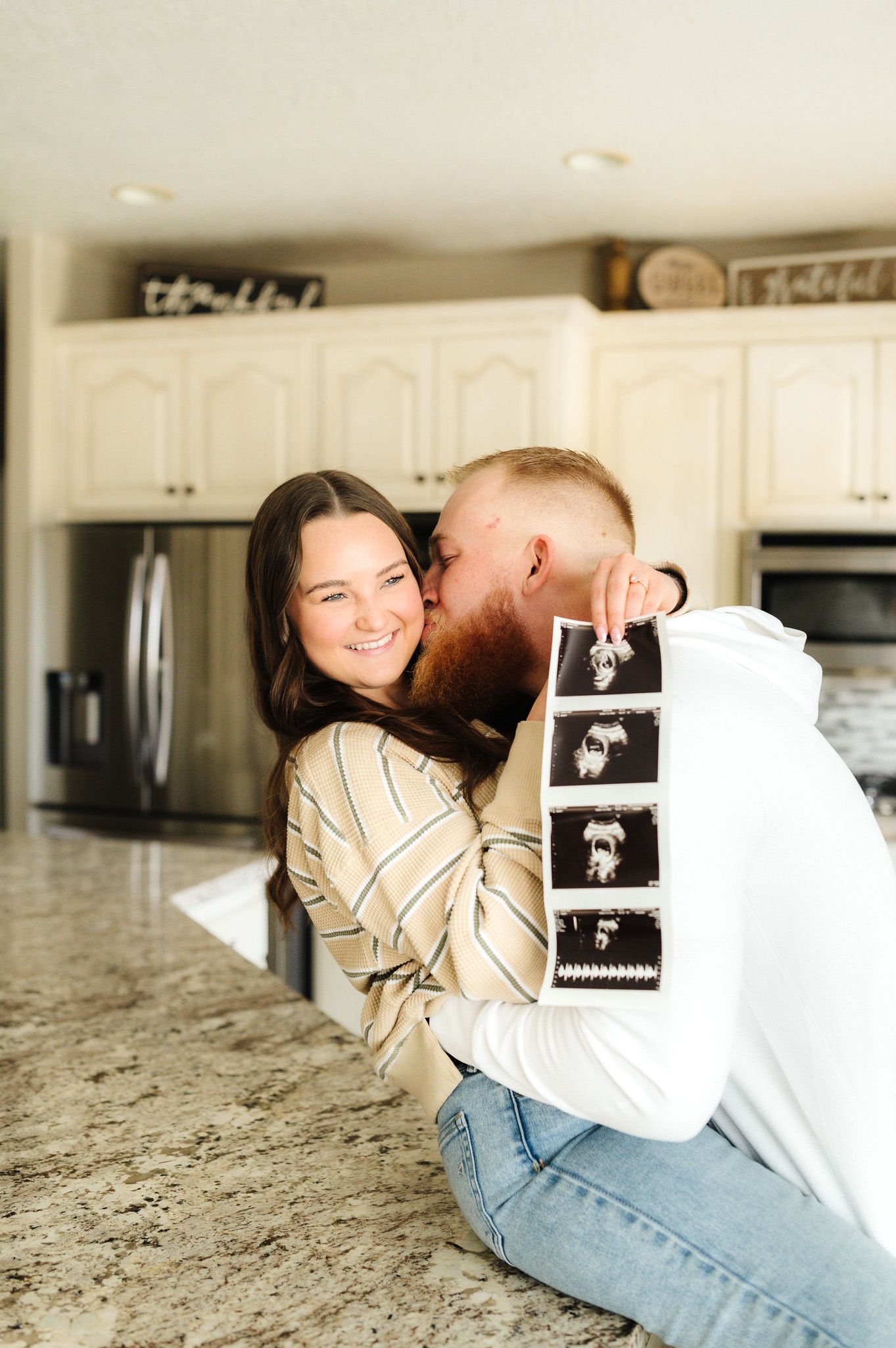 Couple sharing tender moments on kitchen counter while holding ultrasound photos.