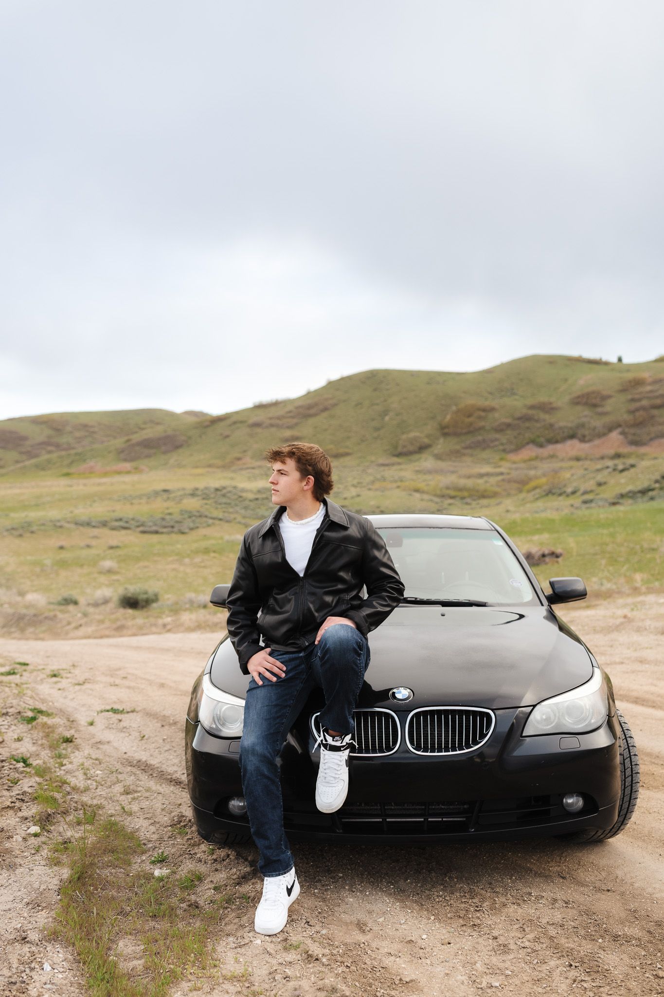 Young man in leather jacket leaning on black BMW sedan on rural dirt road with green hills backdrop.