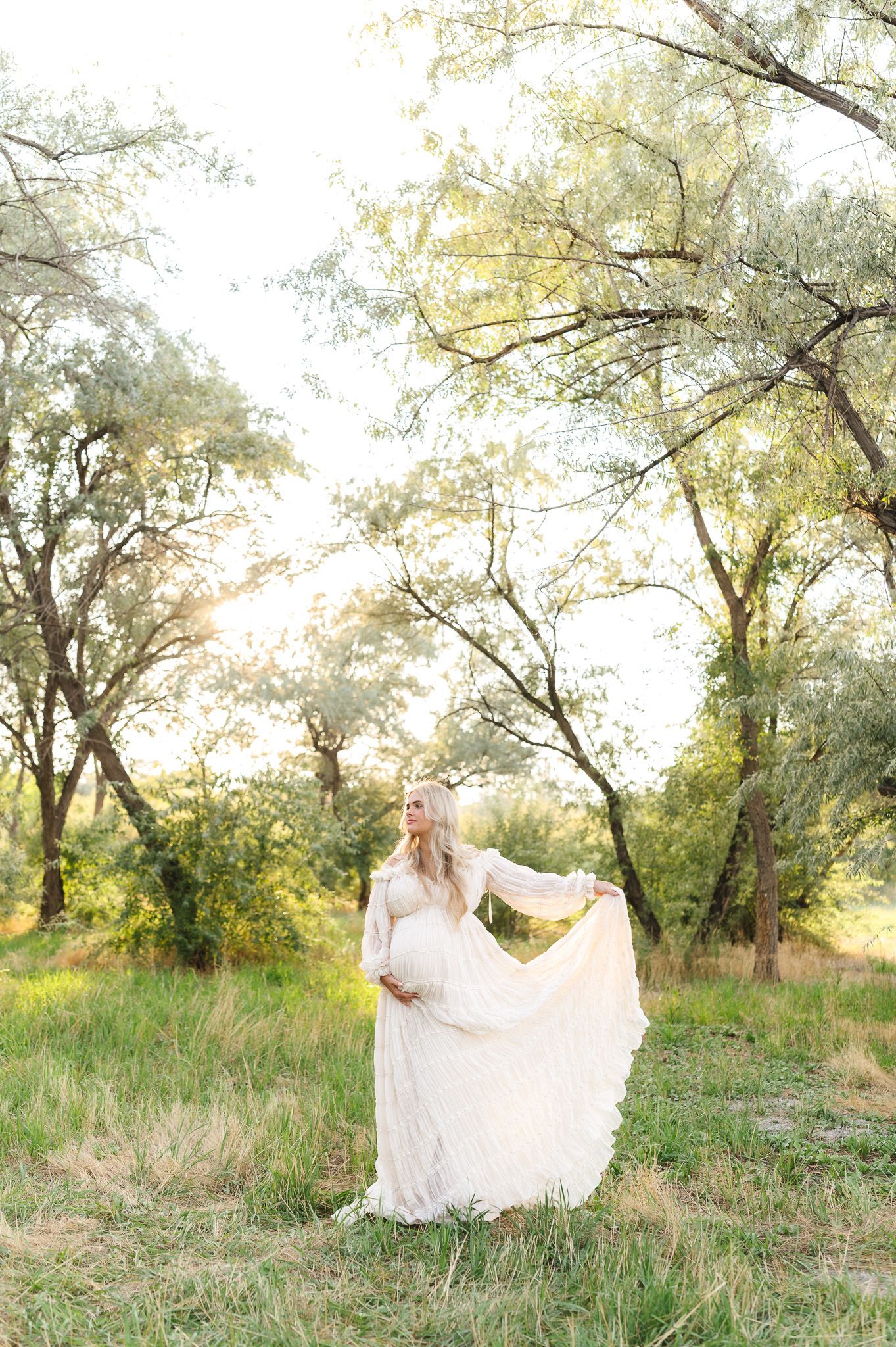 Pregnant woman in white lace gown holding dress hem in sunlit forest clearing with green trees.