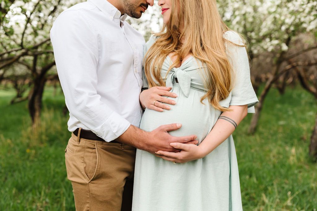 A tender maternity moment captured in a spring orchard with white blossoms in the background.