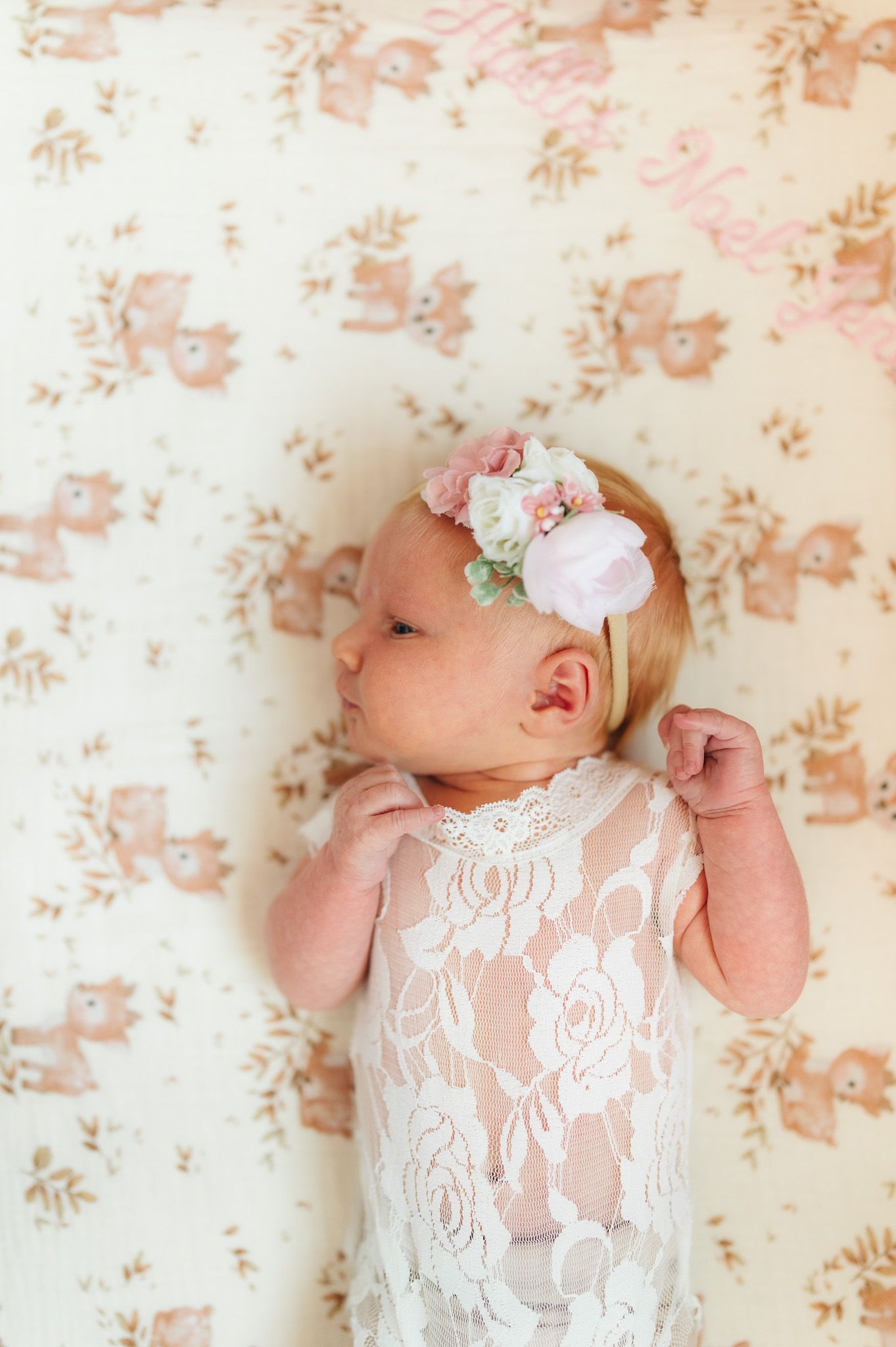 Newborn baby girl in lace romper and floral headband lying on a soft bear-print blanket.