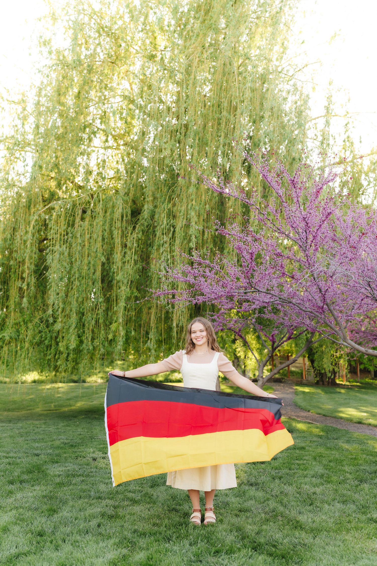 A person in a white dress holds a German flag in front of a blooming cherry tree and weeping willow trees.