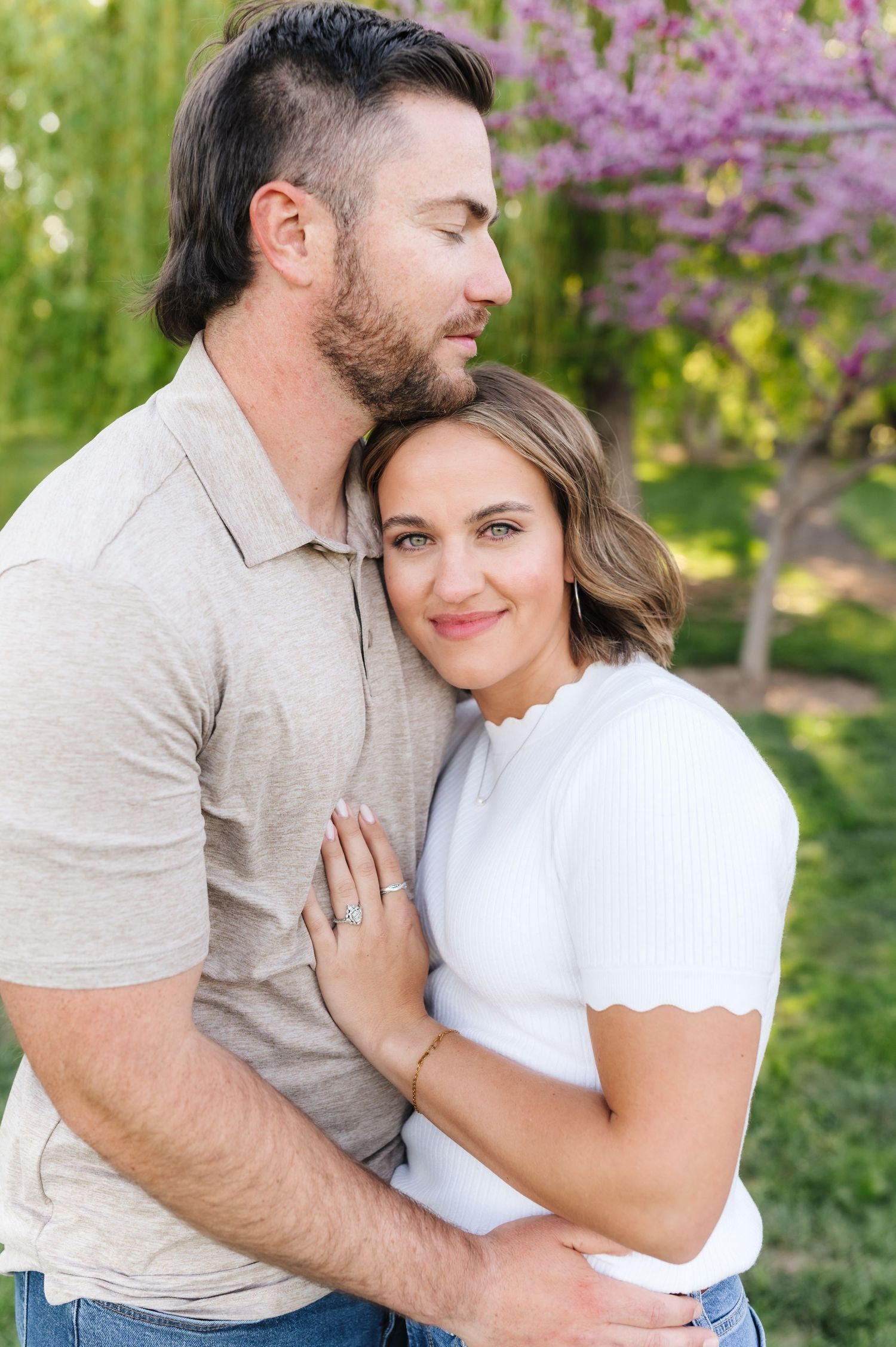 A romantic couple embraces on a spring day with blooming redbud trees and green grass in the background.