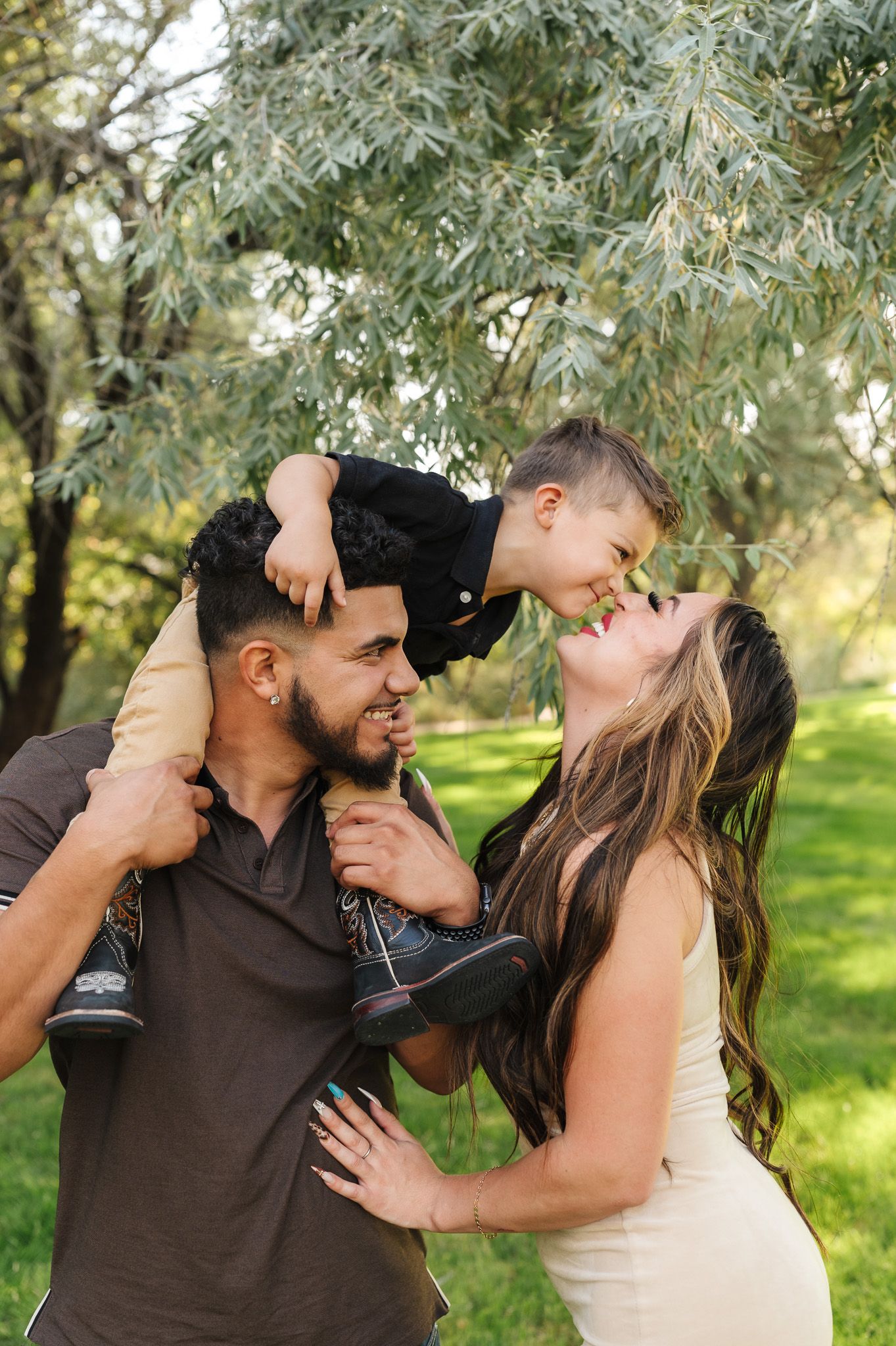 A joyful family moment outdoors as a father lifts his toddler son onto his shoulders while the mother laughs up at the child.