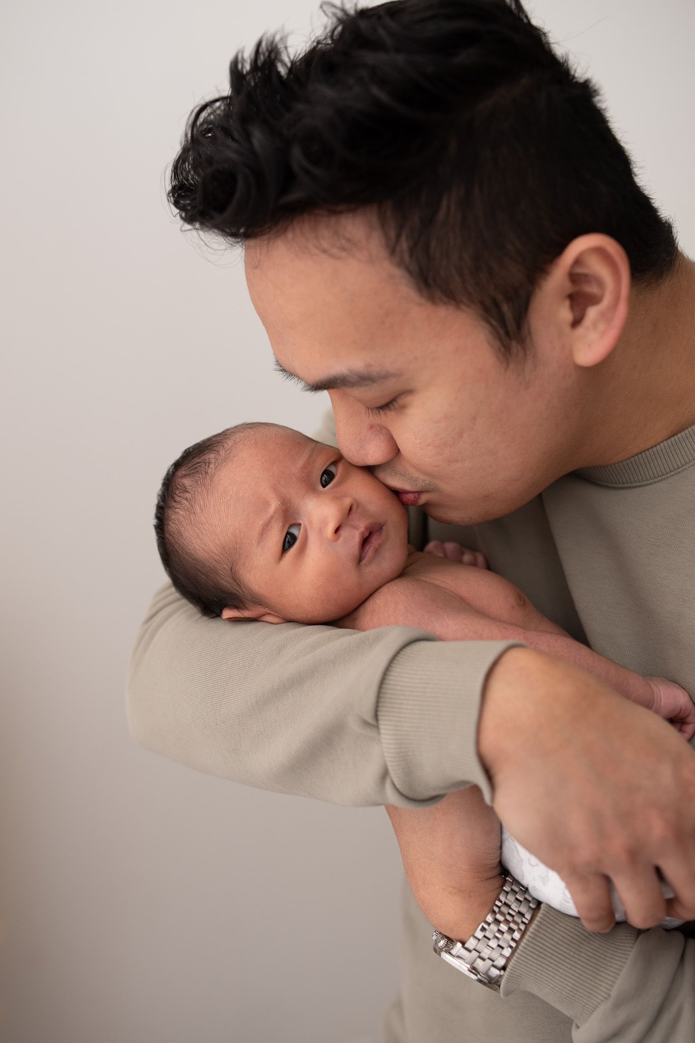 A father tenderly kisses his newborn baby while cradling the infant in his arms against a light background.