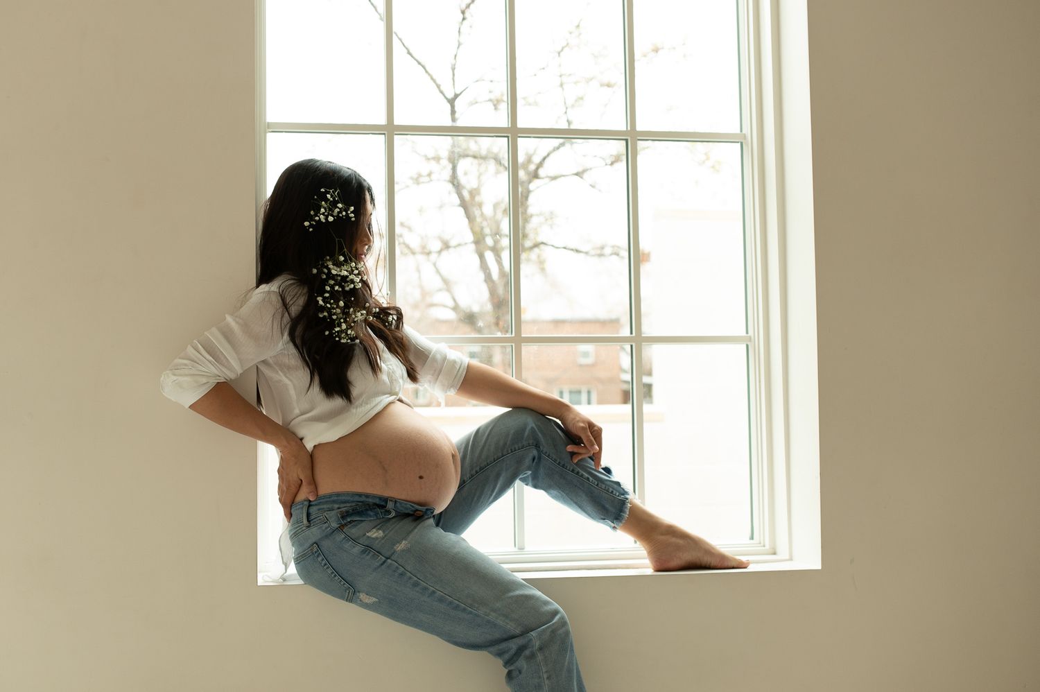 Pregnant woman in white crop top and jeans sitting on window sill with flowers in hair, soft natural light.