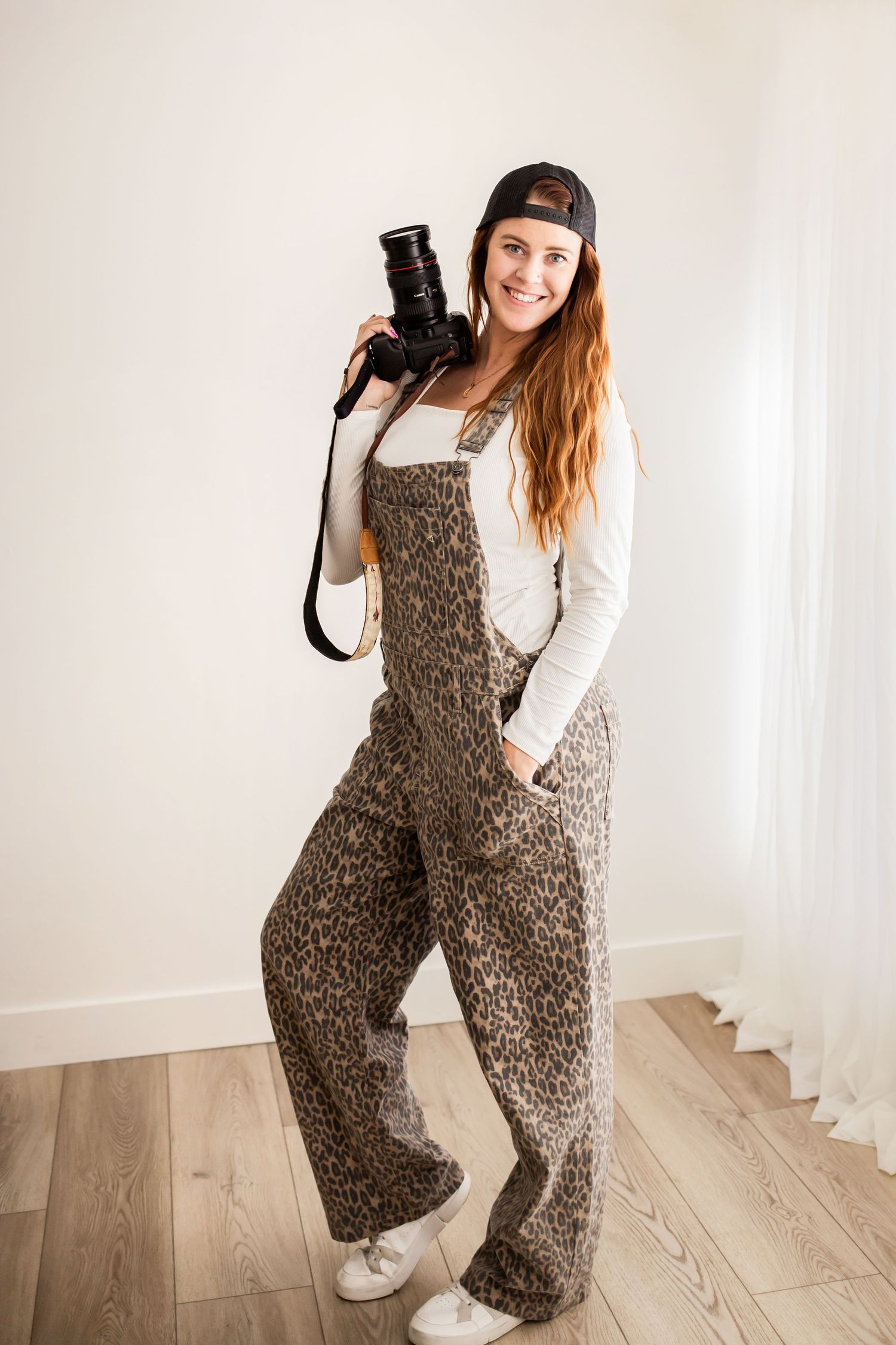 Person wearing leopard print overalls and white sweater poses playfully against white wall.