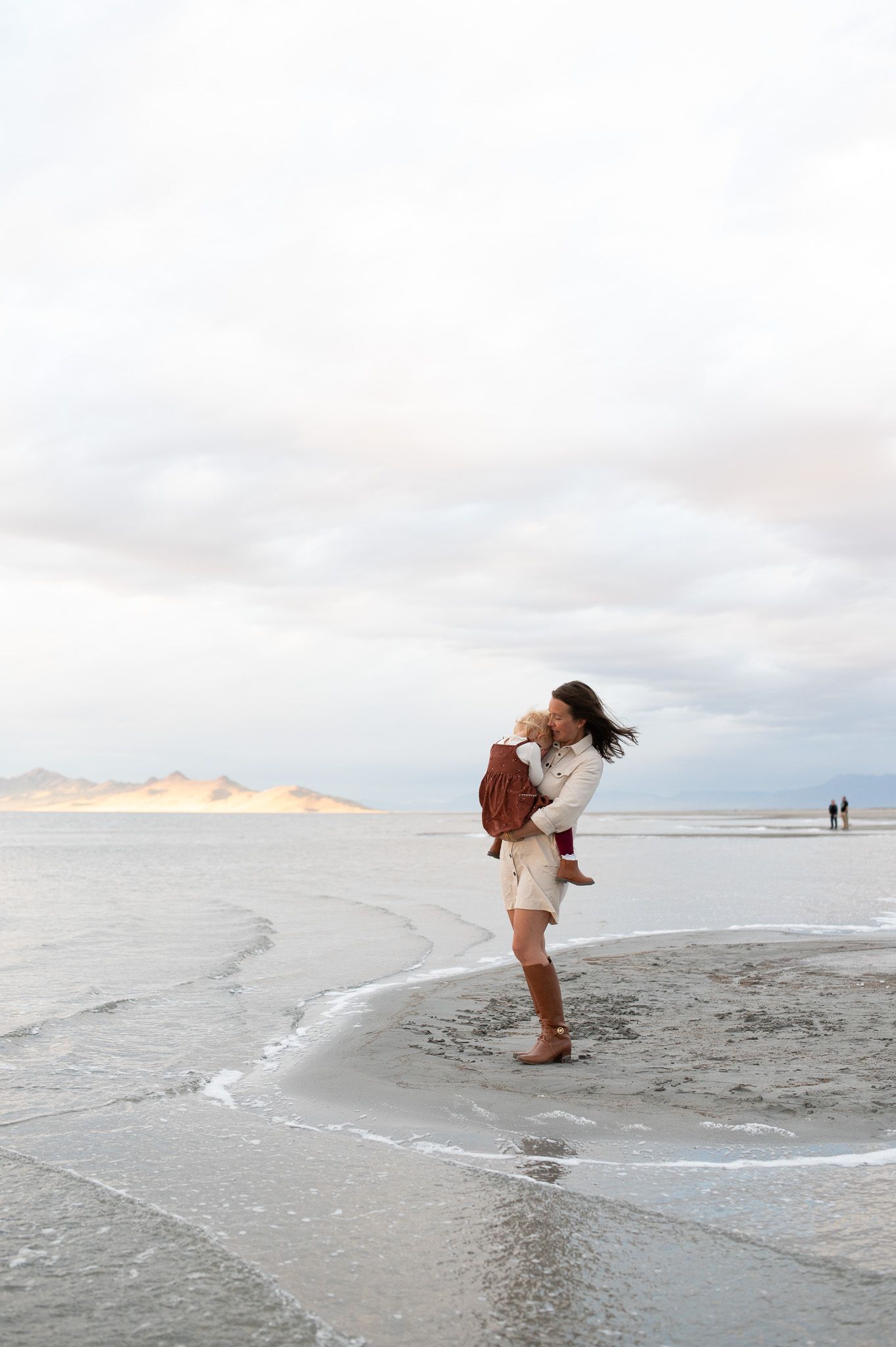 Mother lifting and kissing her baby while standing barefoot on a serene beach with calm waves and cloudy sky.