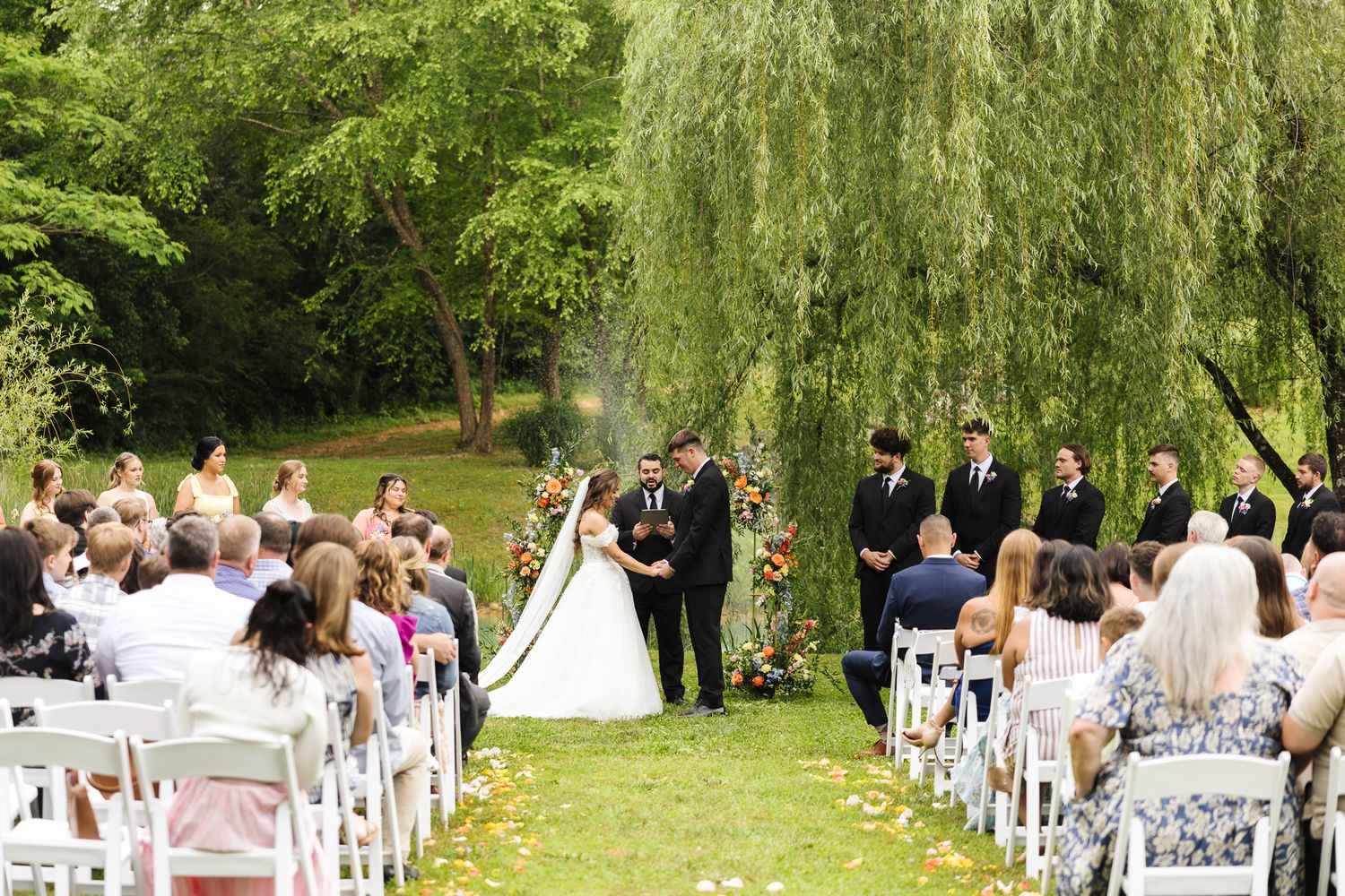 Outdoor wedding ceremony under a willow tree with bride and groom at floral arch, guests seated in white chairs on green lawn.