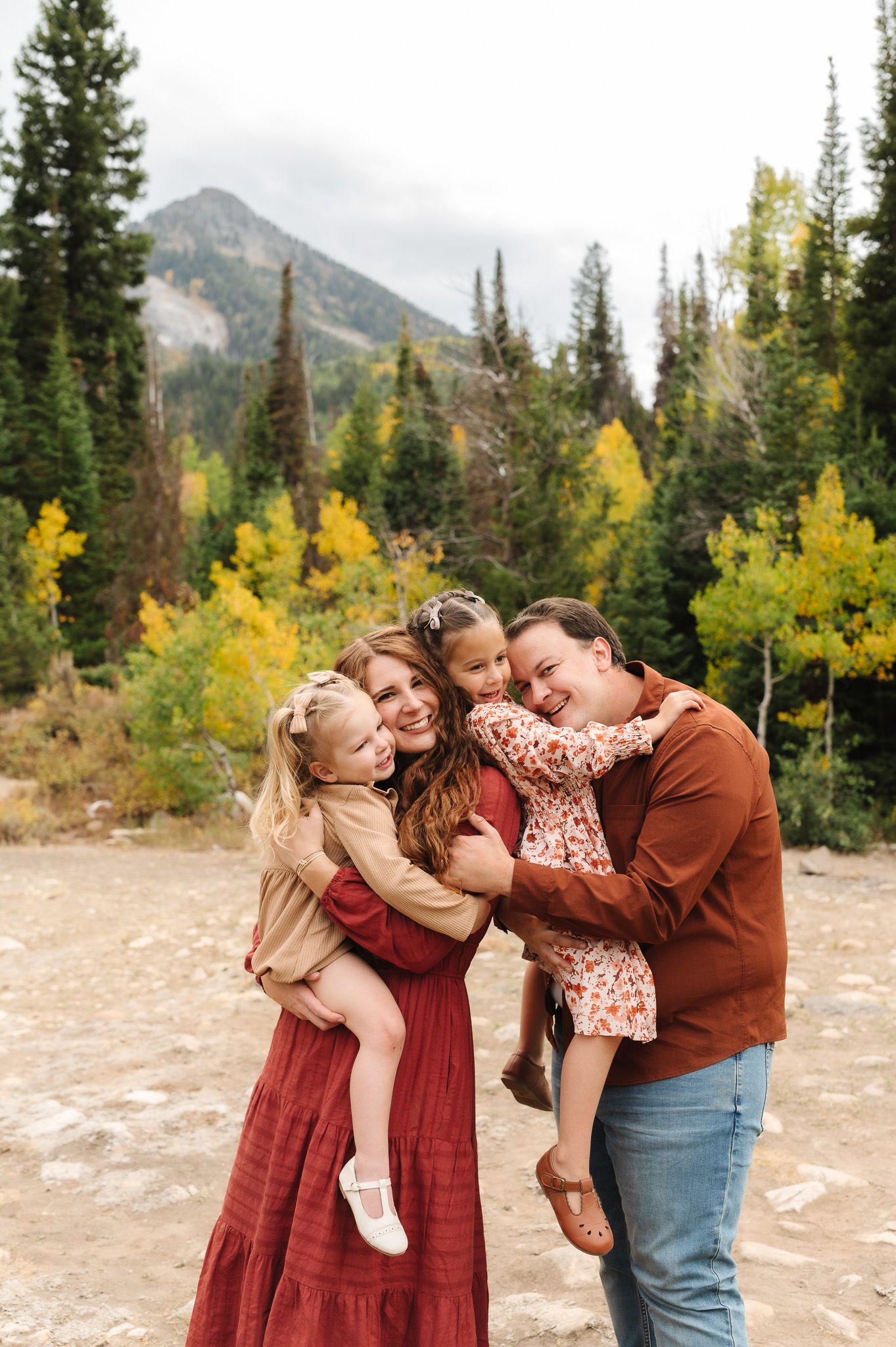 Happy family of four embracing outdoors with autumn trees and mountain backdrop during fall season.