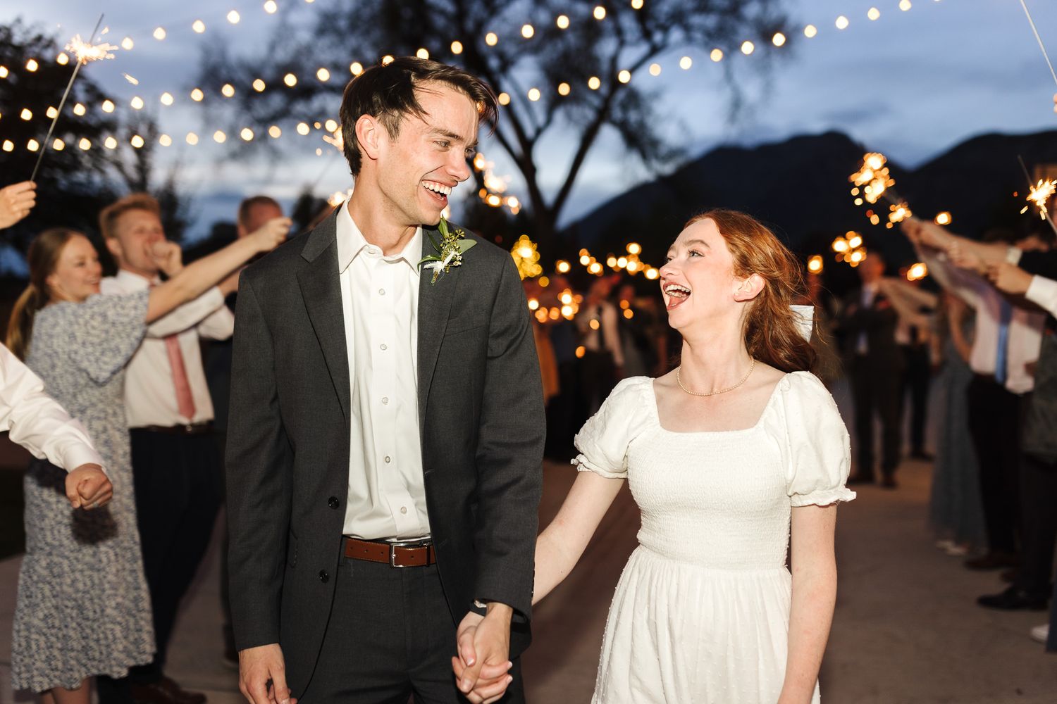 Happy couple walks hand-in-hand through sparkler exit at outdoor wedding reception under string lights at dusk.