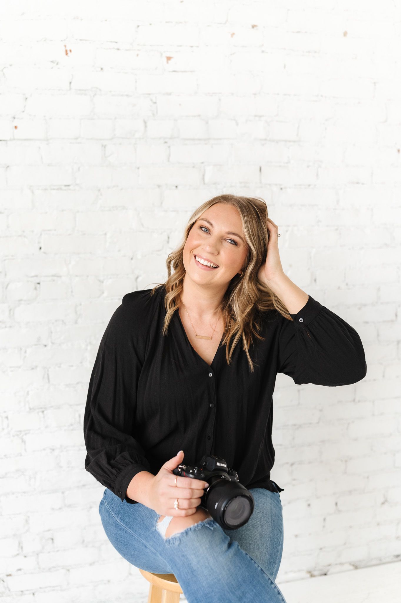 Professional photographer in black top holding camera while sitting casually against white brick wall.