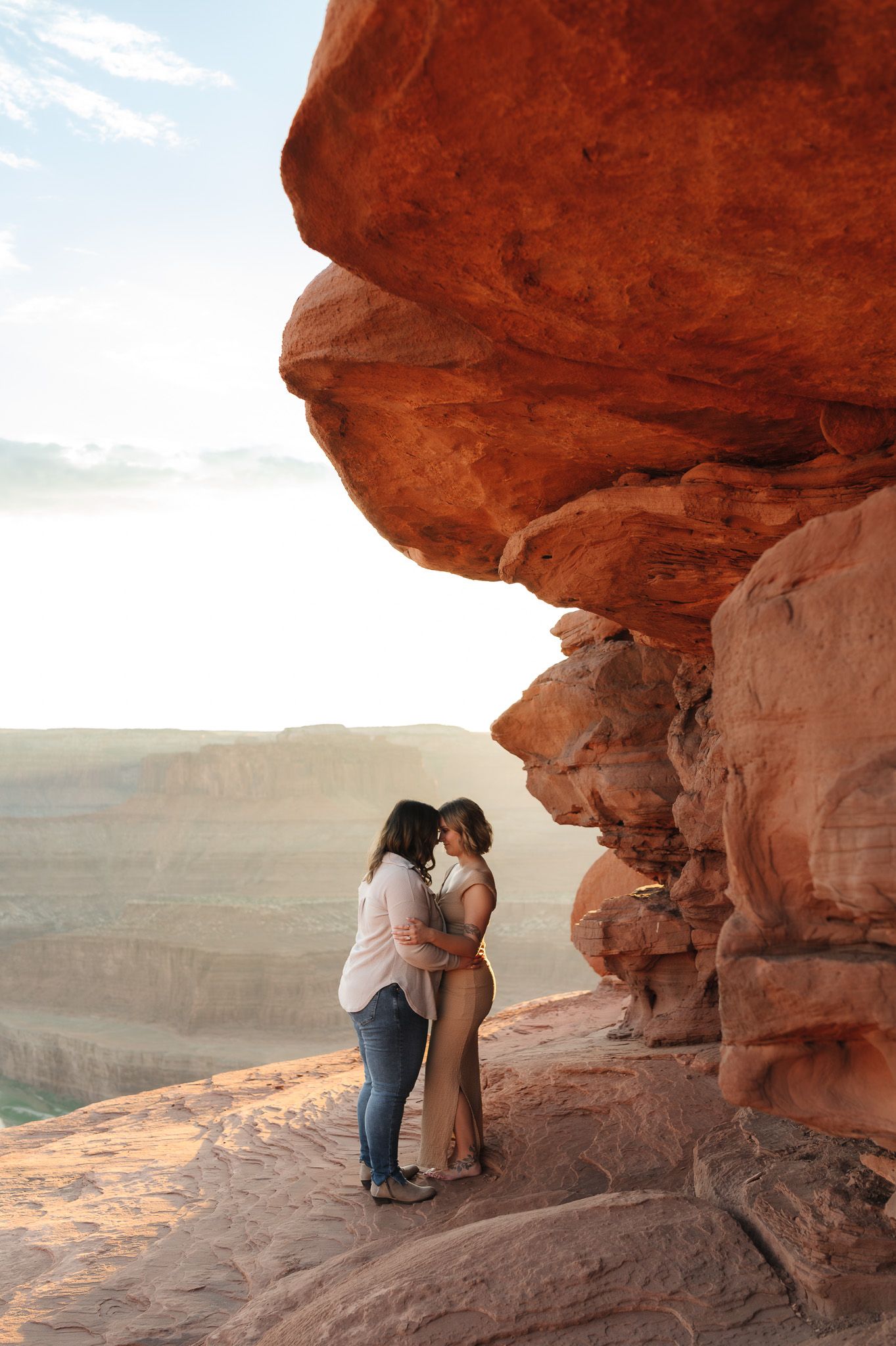 Couple embracing under red rock formation at canyon overlook during golden hour in the American Southwest.