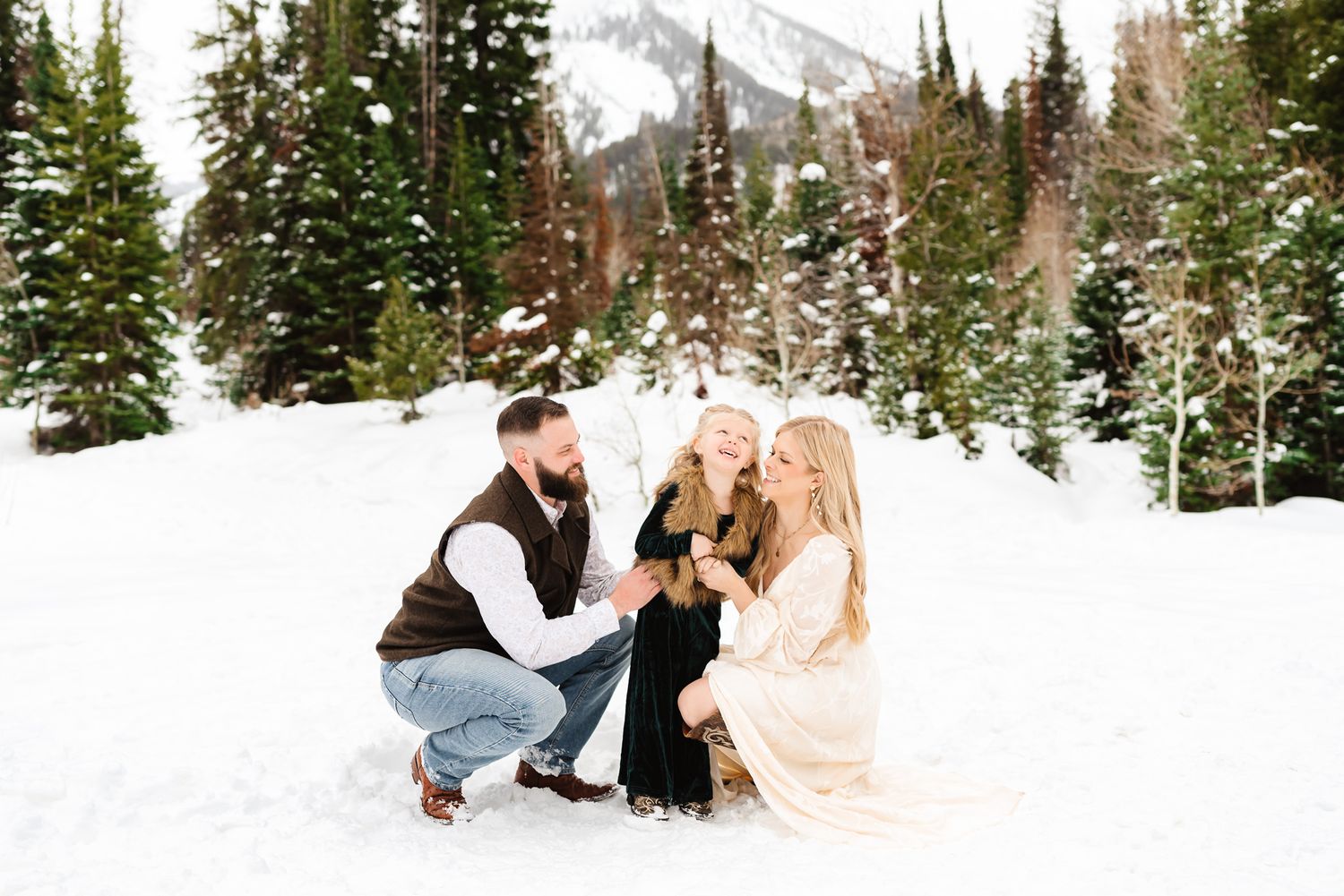 Family members playing and posing in the snow with mountains and evergreen trees in the background during Jordan Pines winter family photos