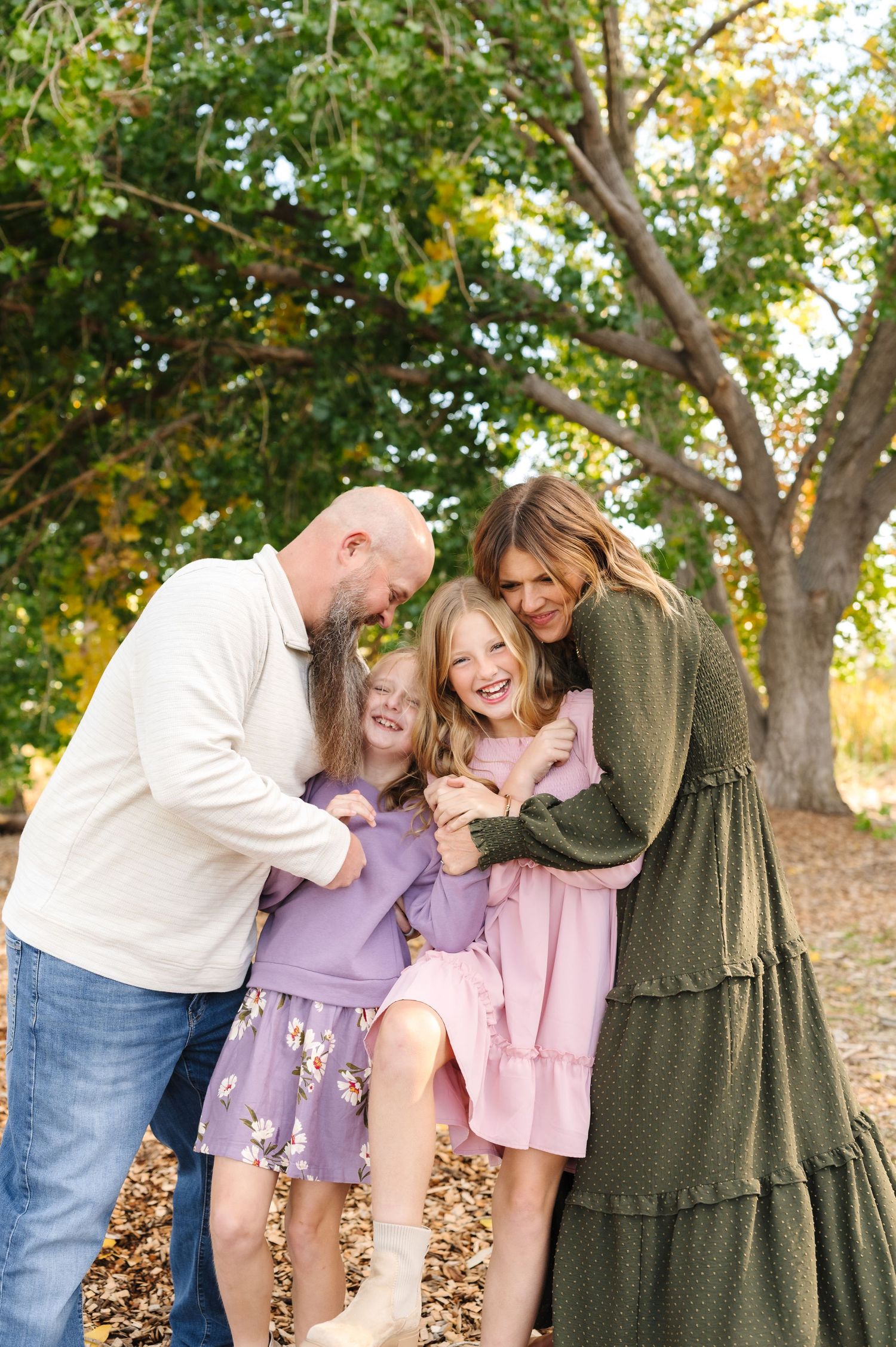 Family of four laughing together outdoors in fall, parents hugging two young girls in colorful dresses among autumn leaves.