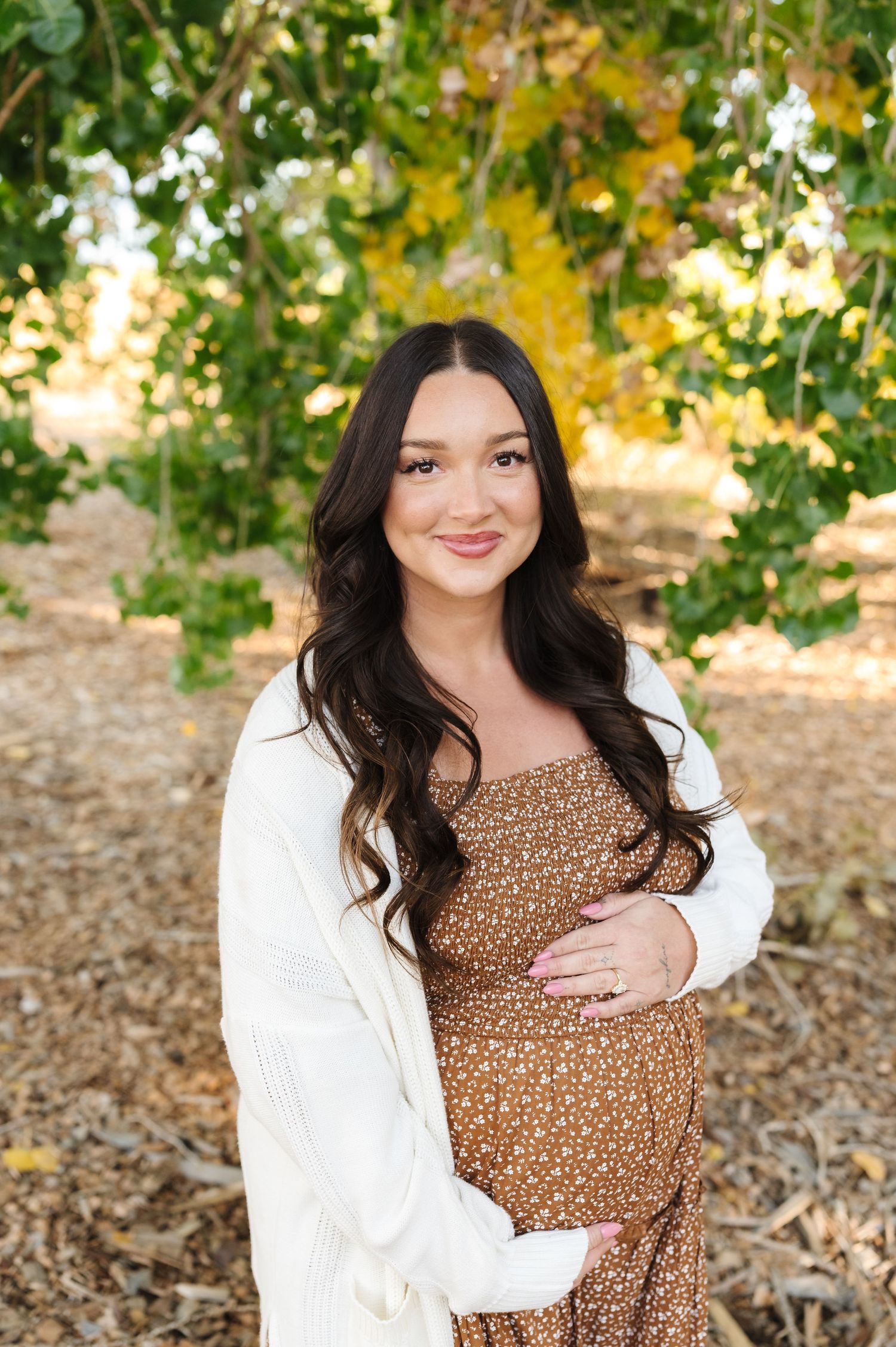 Pregnant woman in brown floral dress and white cardigan smiling outdoors among green and yellow autumn trees.