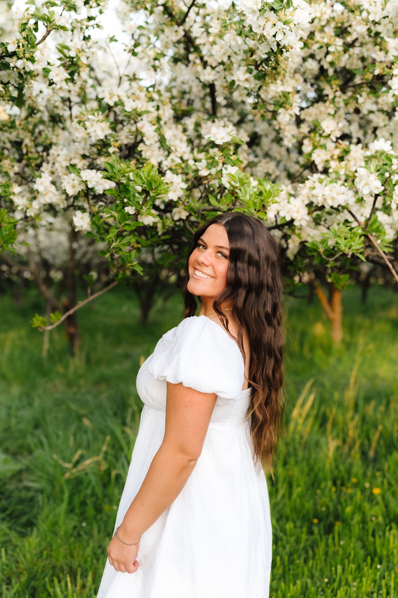 A person in a white dress poses joyfully among blooming apple trees in a spring orchard.