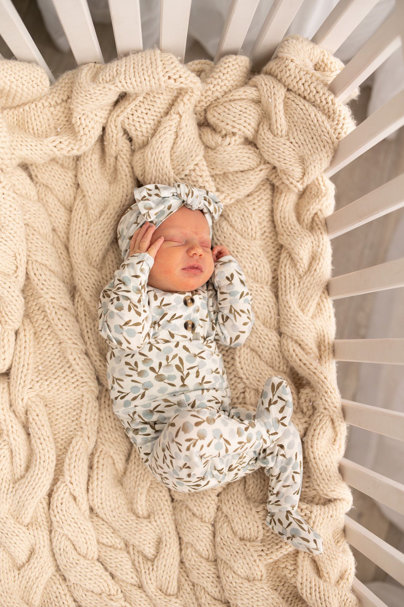 Newborn baby sleeping peacefully in a crib on a chunky knit cream blanket, wearing a floral footie onesie and matching bow headband.