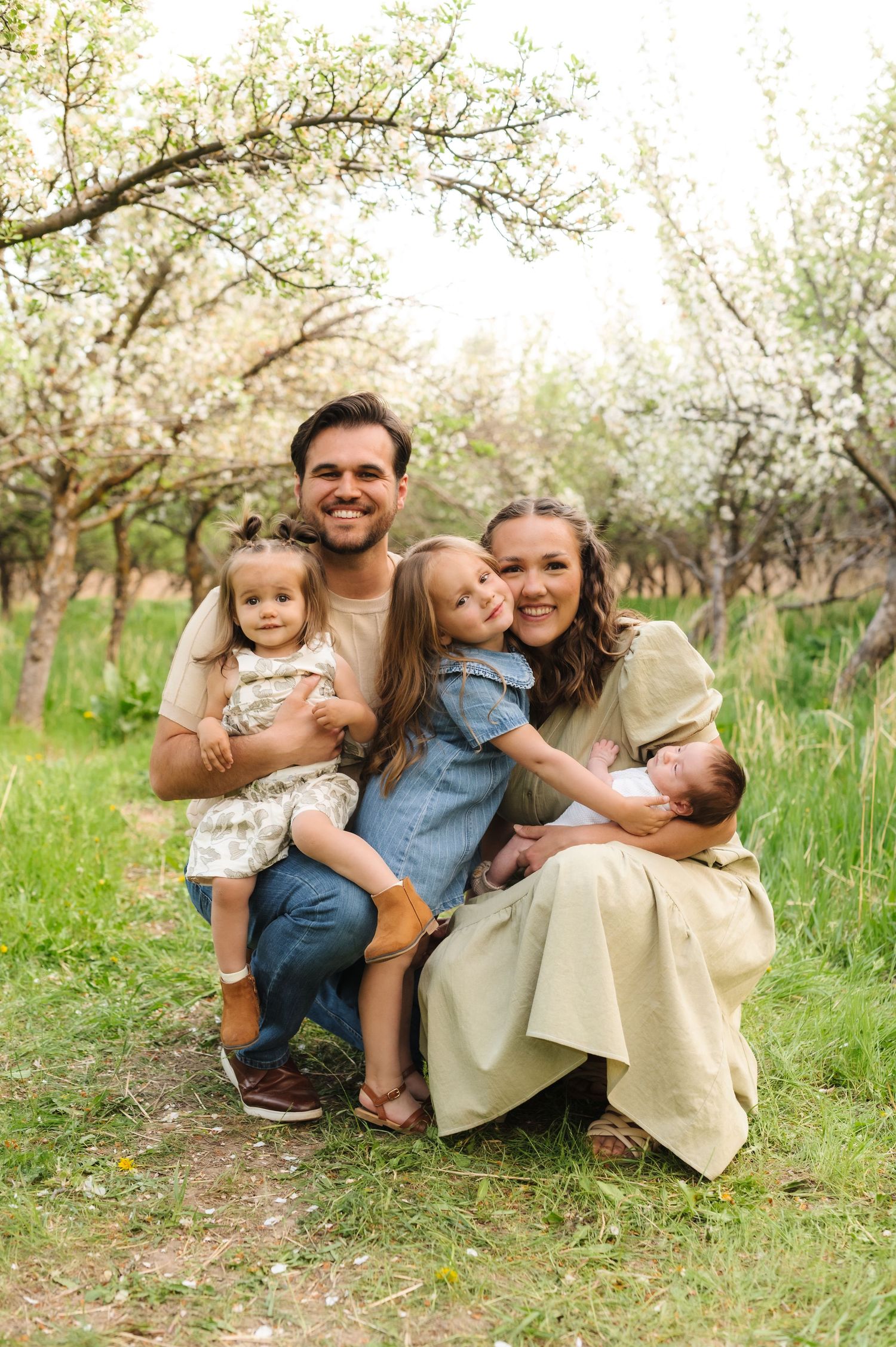 Family laughing together while sitting in blooming spring orchard with white blossoms and green grass.