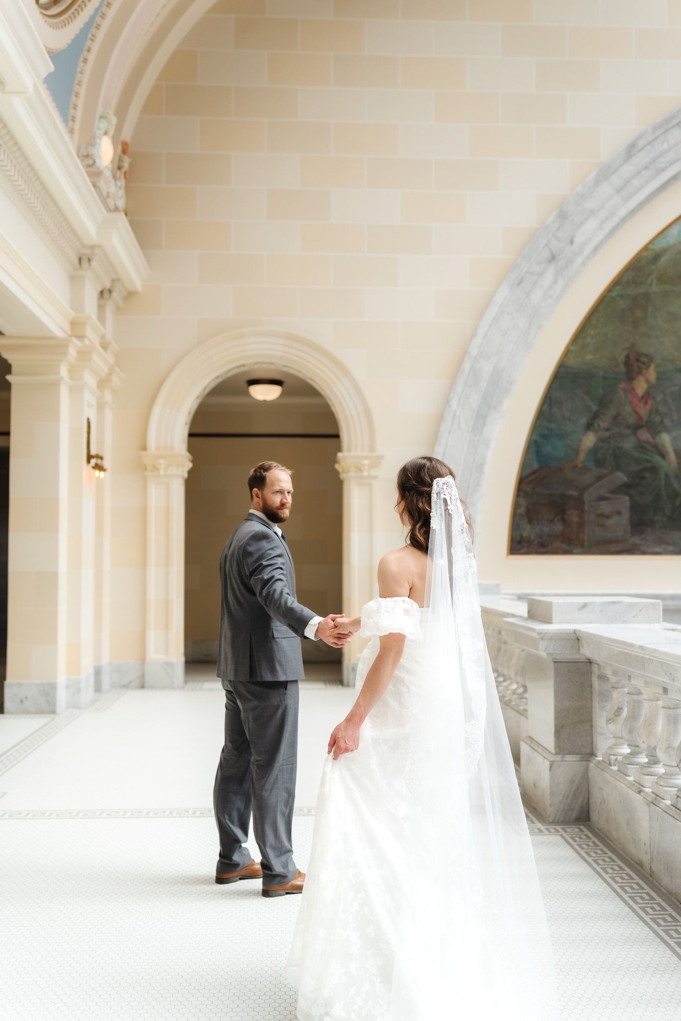 Bride and groom hold hands in an elegant marble hallway with arched columns and a classical mural during their wedding.