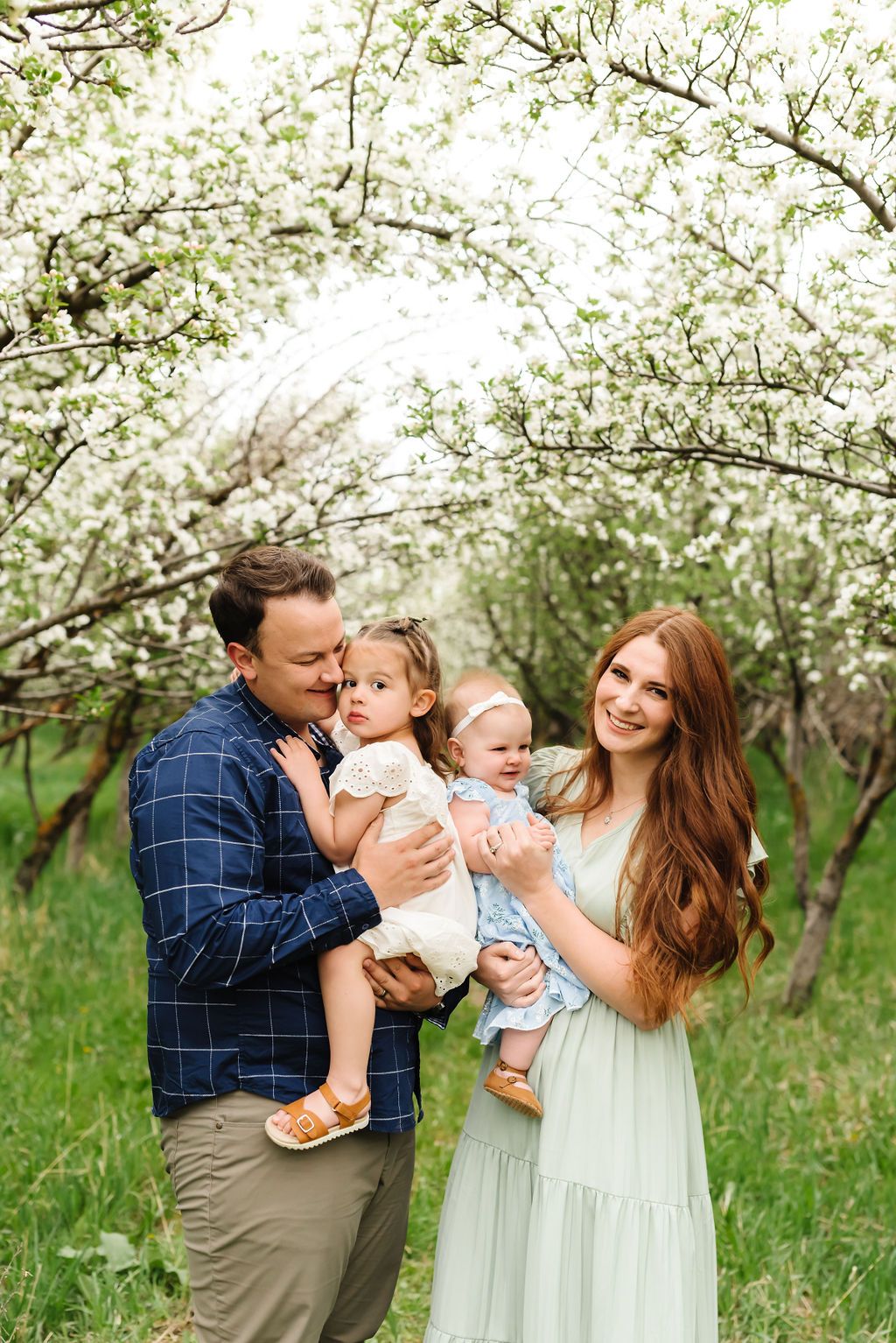A family embraces together in a serene orchard setting with blooming white spring blossoms overhead.