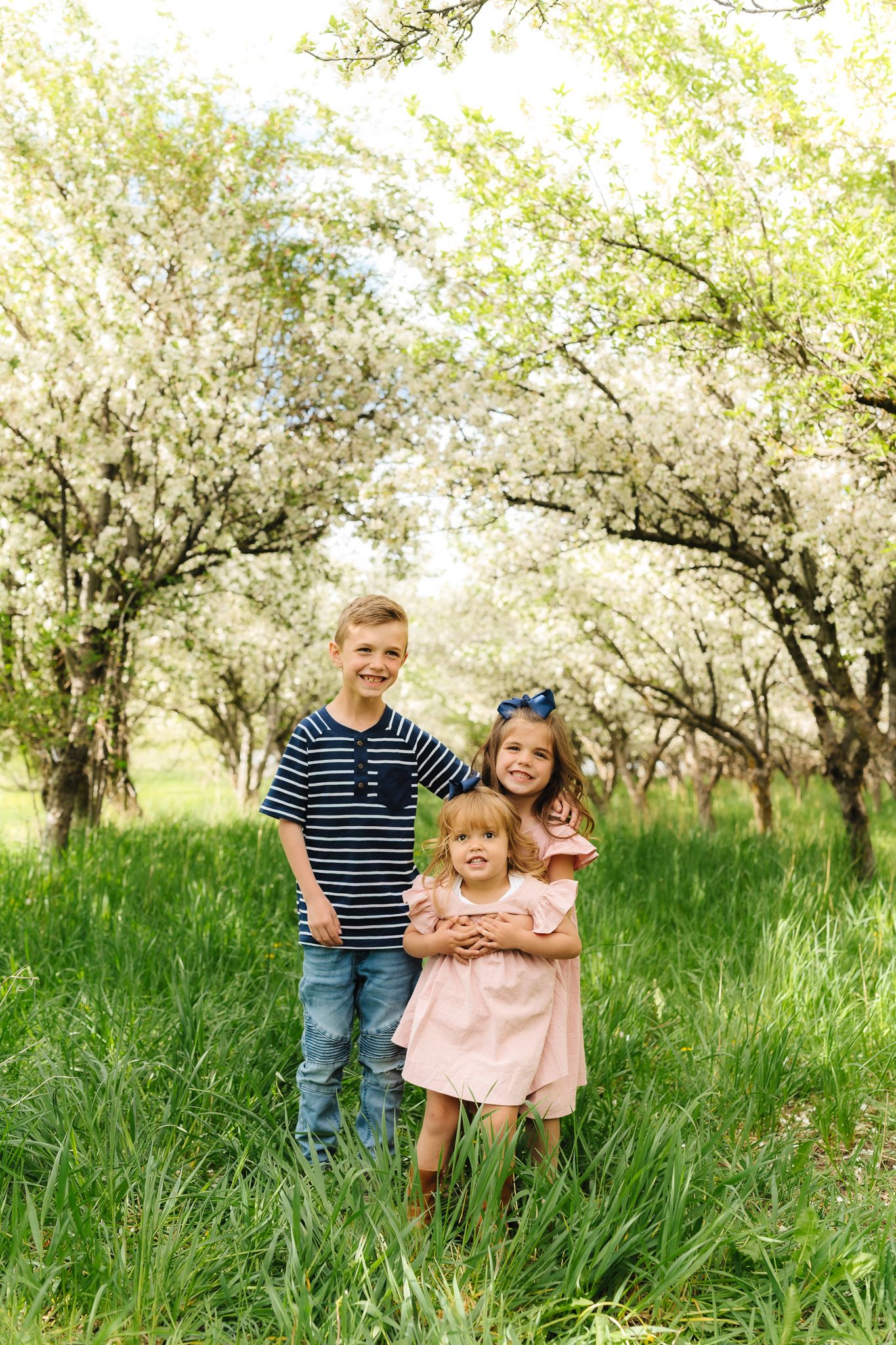 Three children pose together under flowering trees in a spring orchard setting.