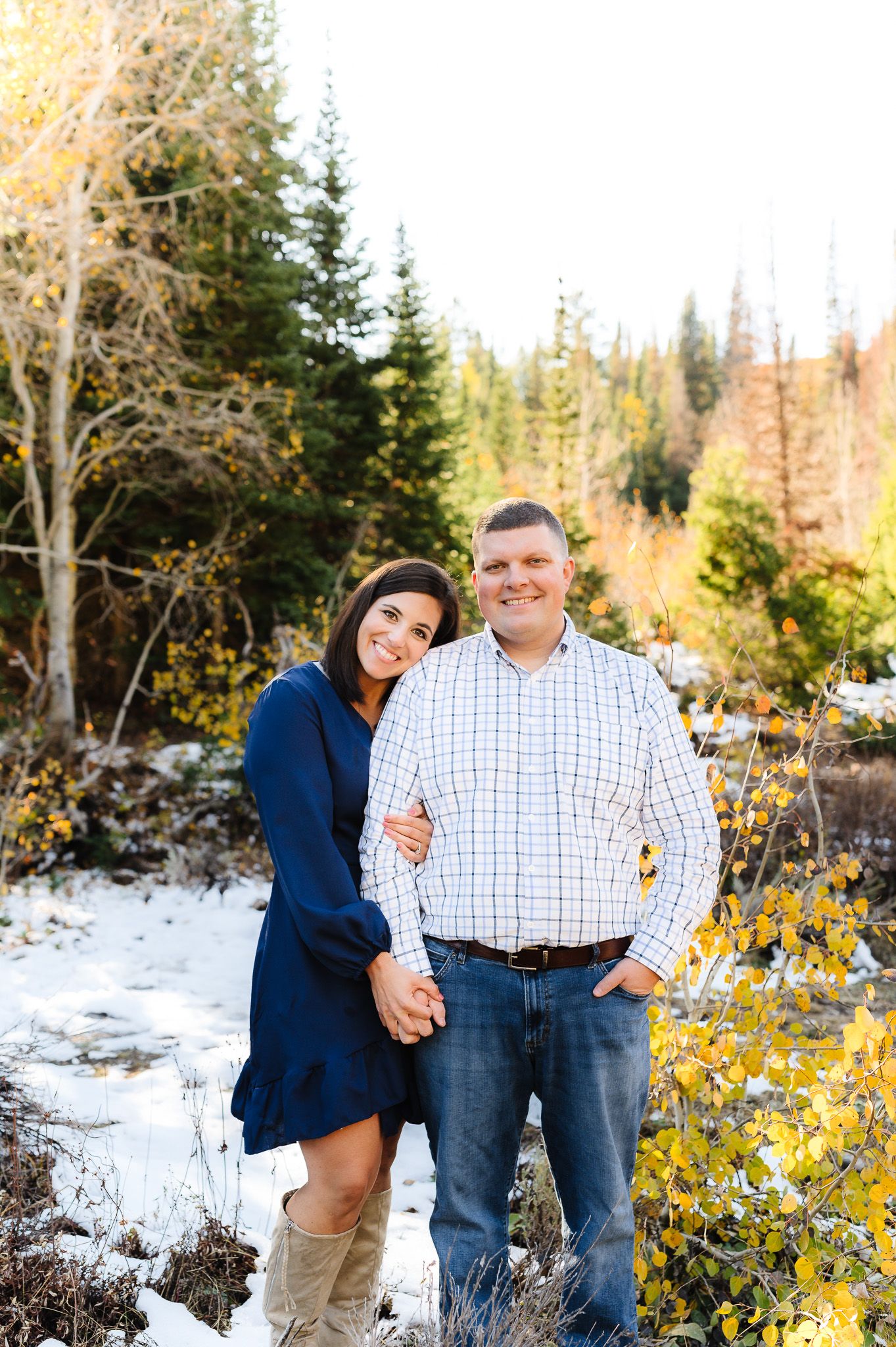 A couple poses together in a snowy mountain setting with evergreen trees and autumn foliage in the background.