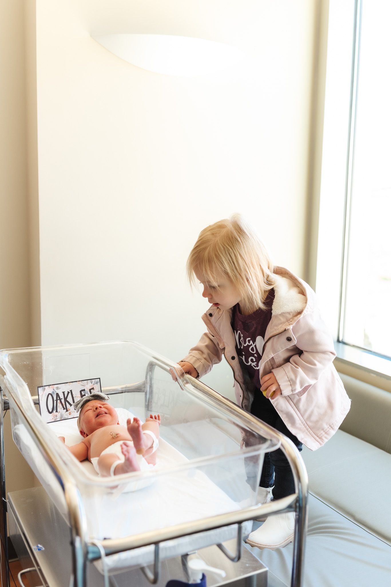 Young blonde girl leaning over hospital bassinet looking at newborn baby named Oaklee in maternity ward.