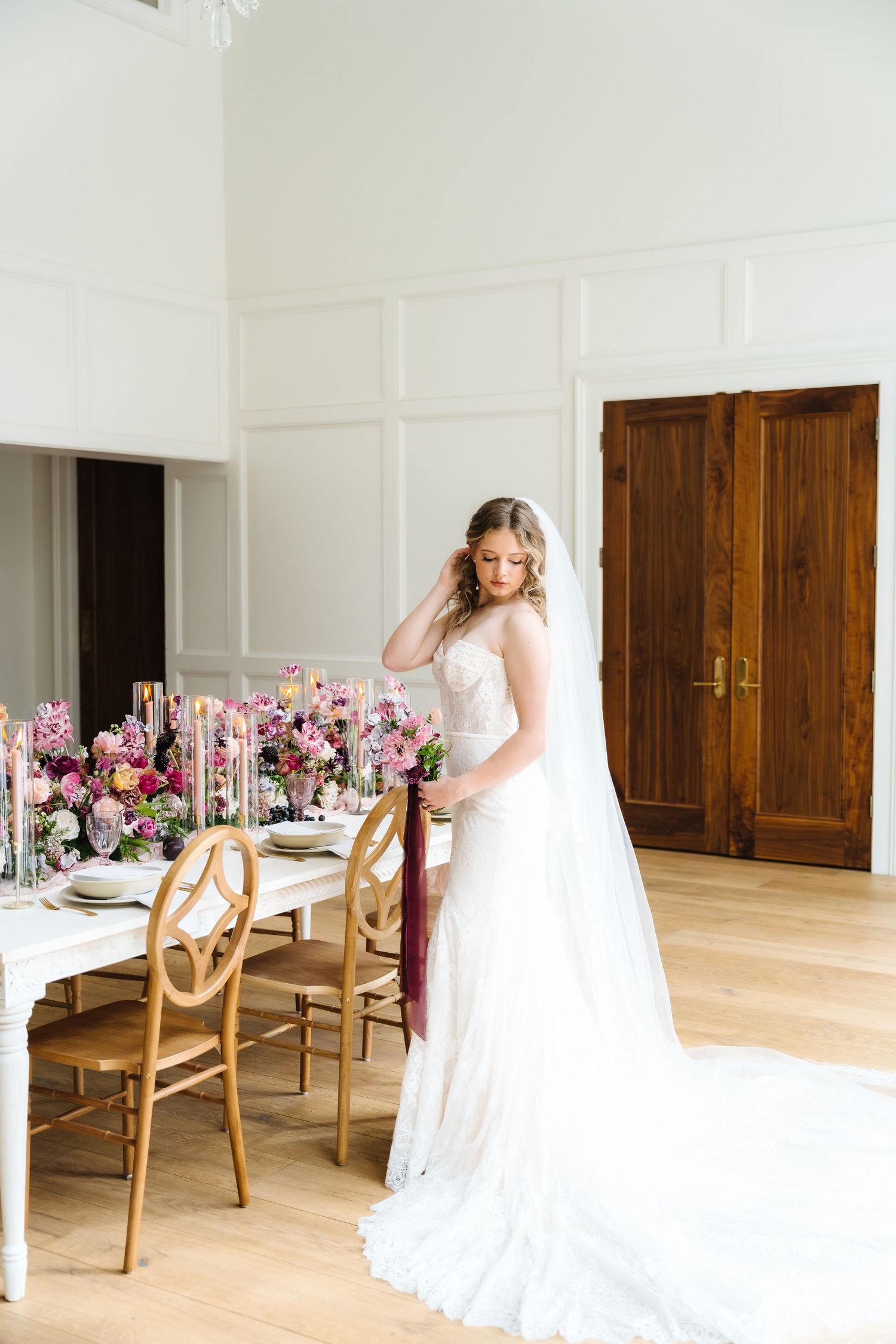 Wedding dress with flowing train in bright venue featuring rustic wooden chairs and pink floral centerpieces.