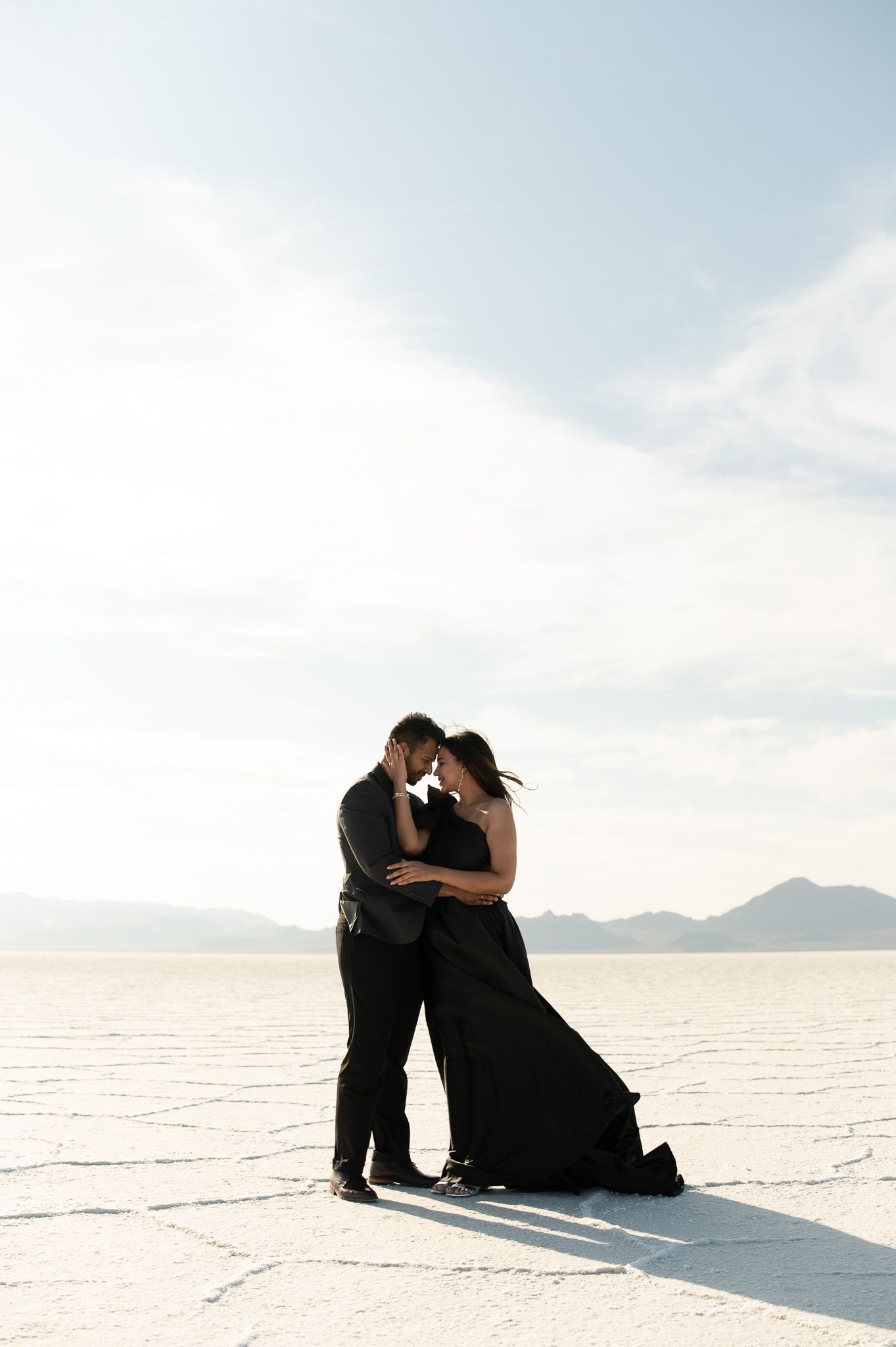 Couple in black attire embracing on the Bonneville Salt Flats with mountains and bright sky behind them.