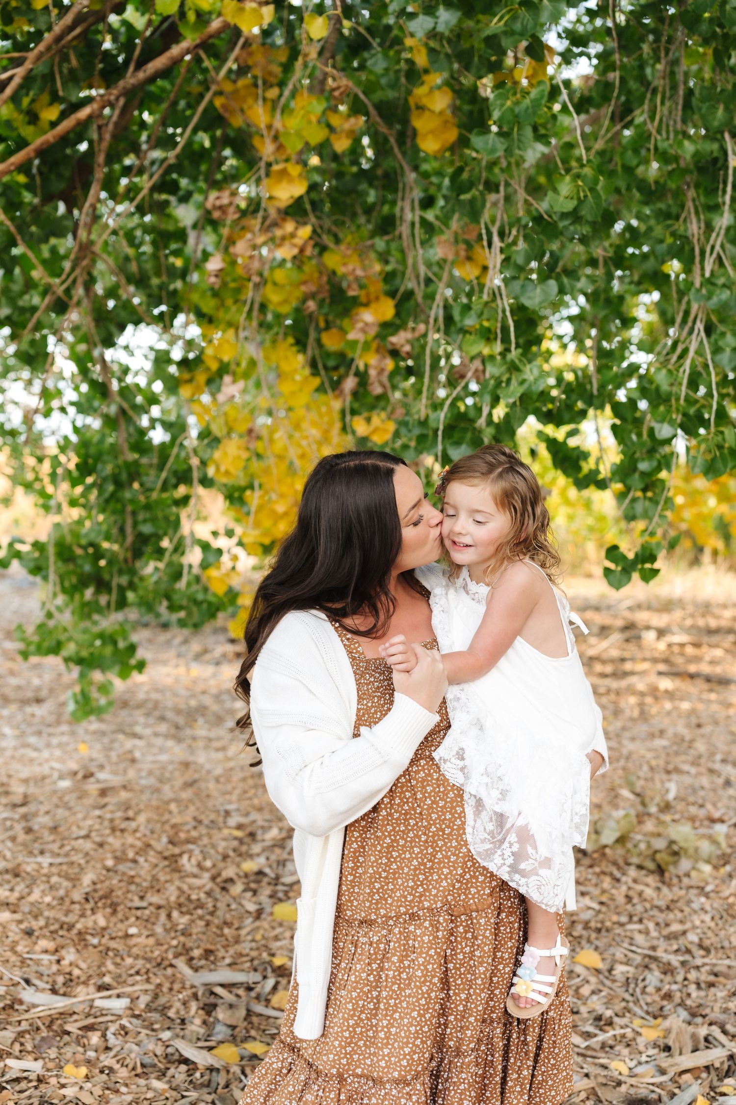 A mother kisses her smiling toddler daughter outdoors under green and yellow foliage in matching white and brown outfits.