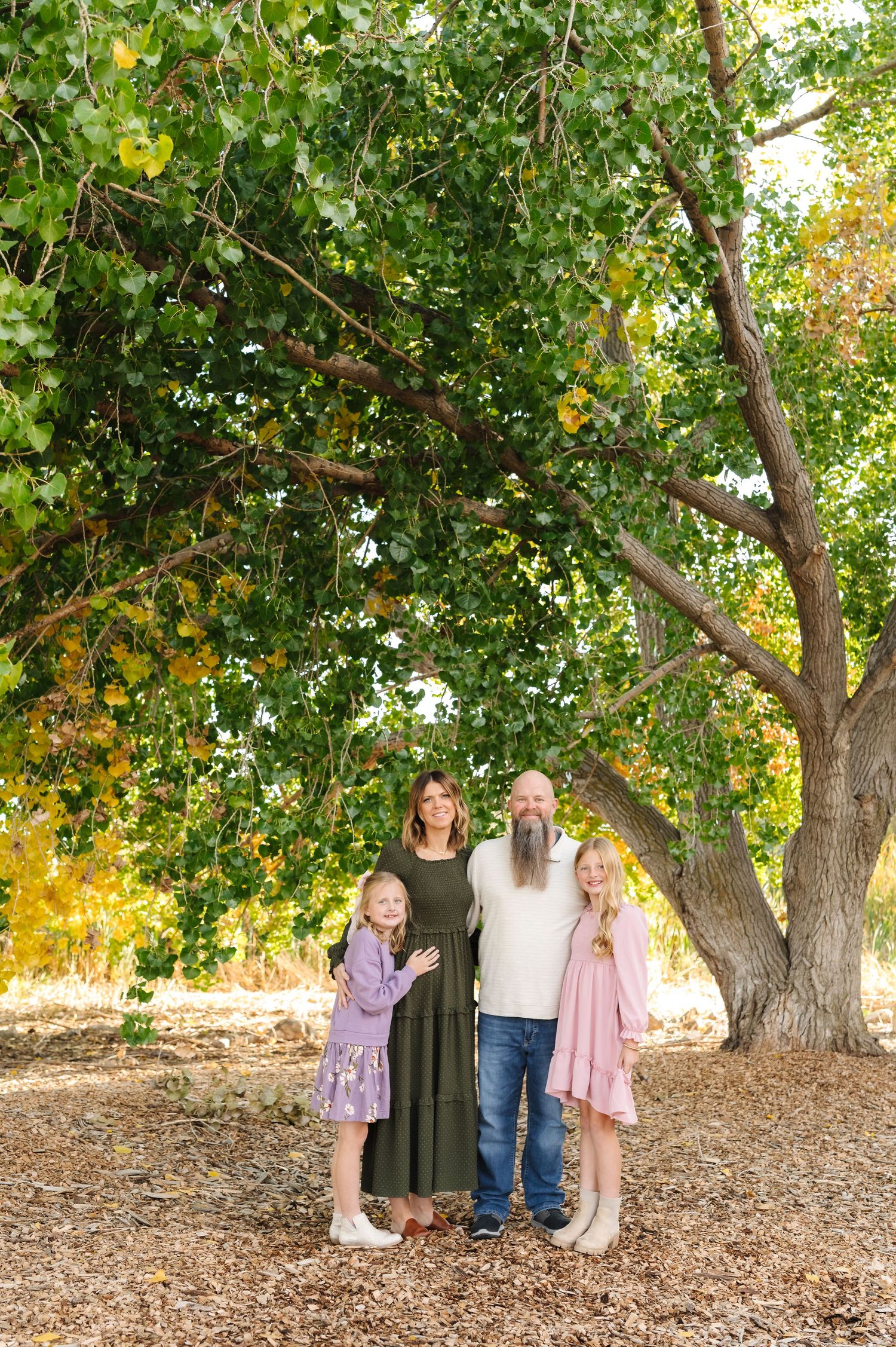 Family of four smiling together under a large green tree on a leaf-covered ground in autumn.