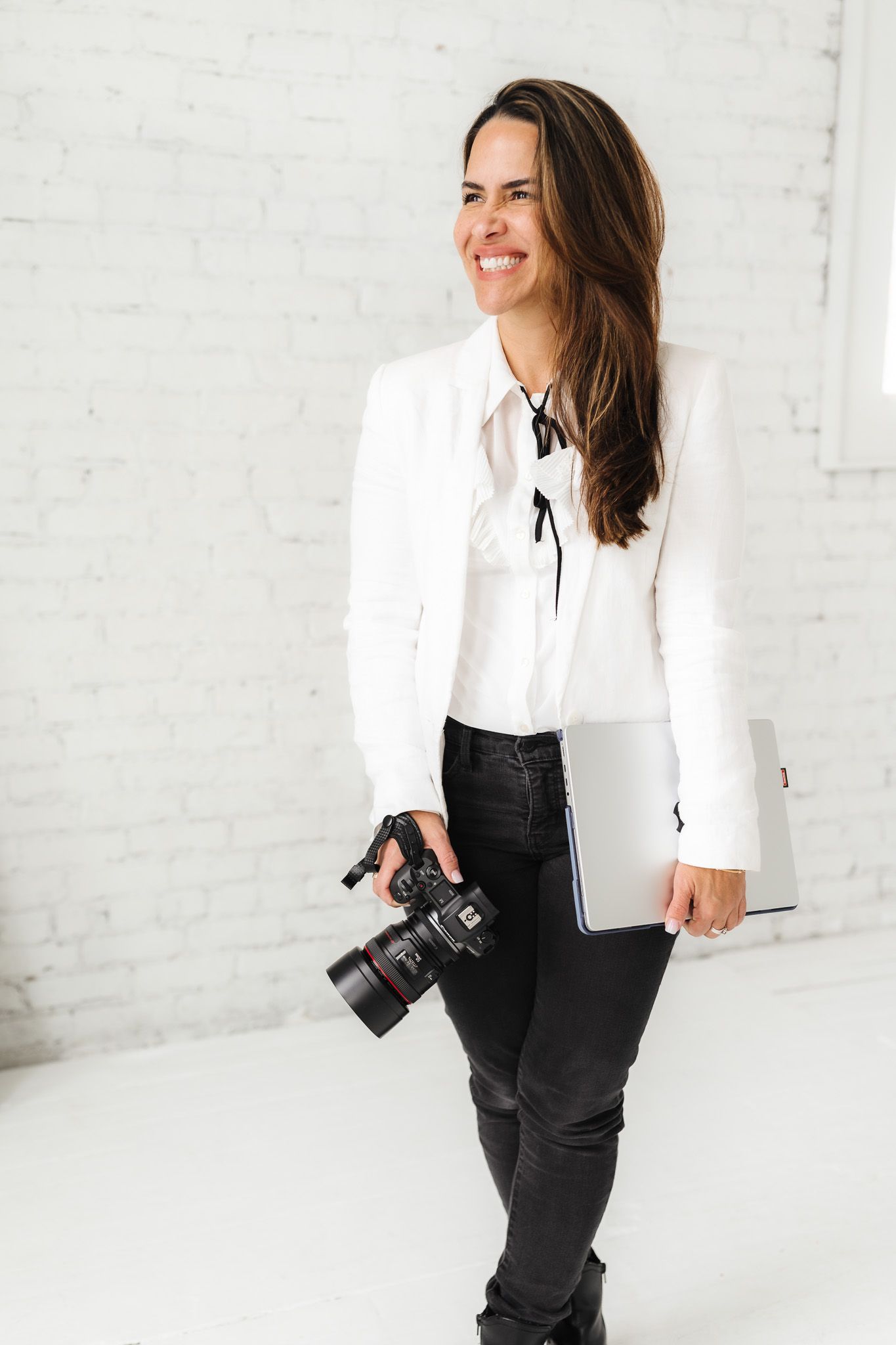 Professional photographer in white blazer and black pants holding camera in bright studio.
