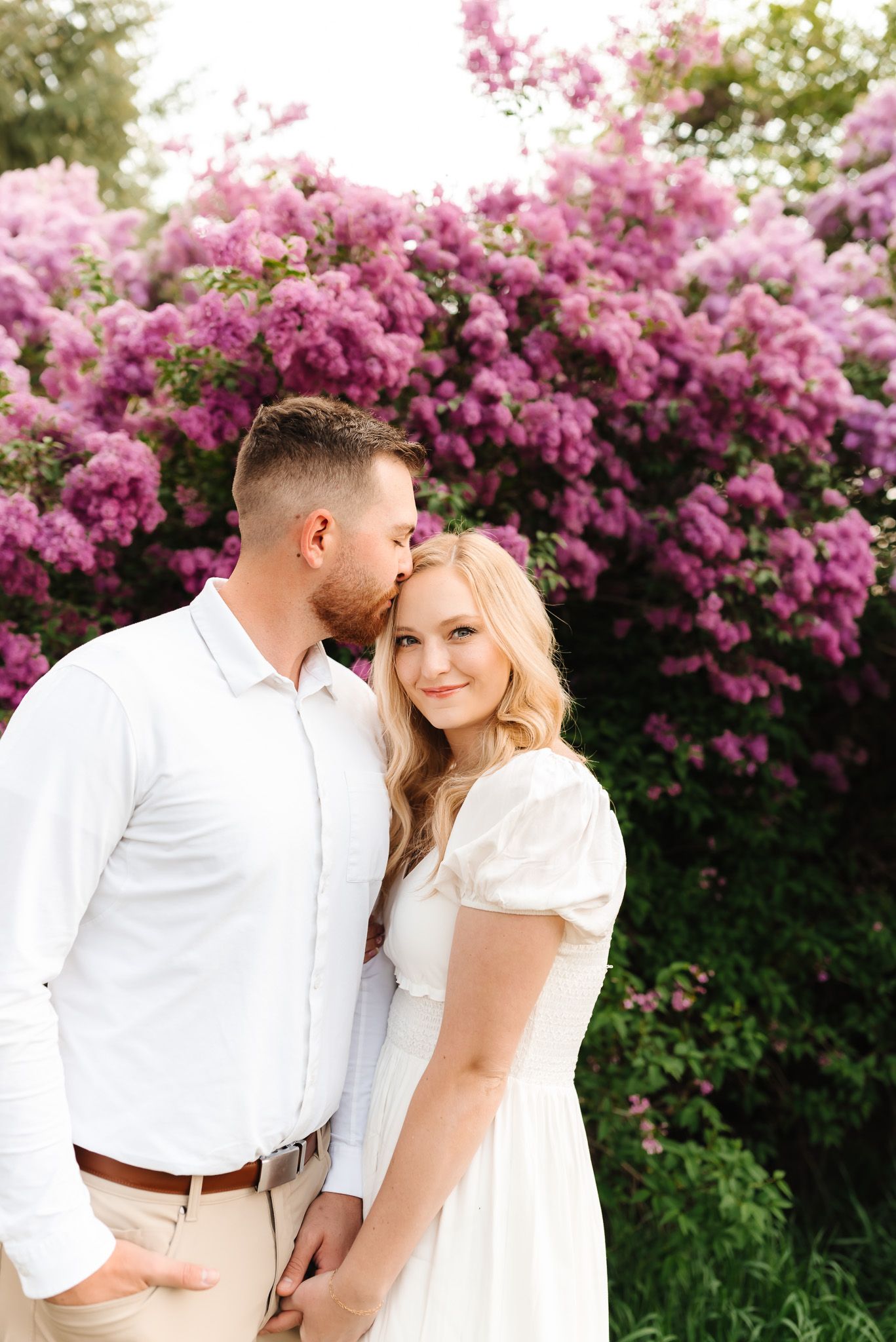 Romantic couple in white outfits pose against vibrant pink flowering bushes.