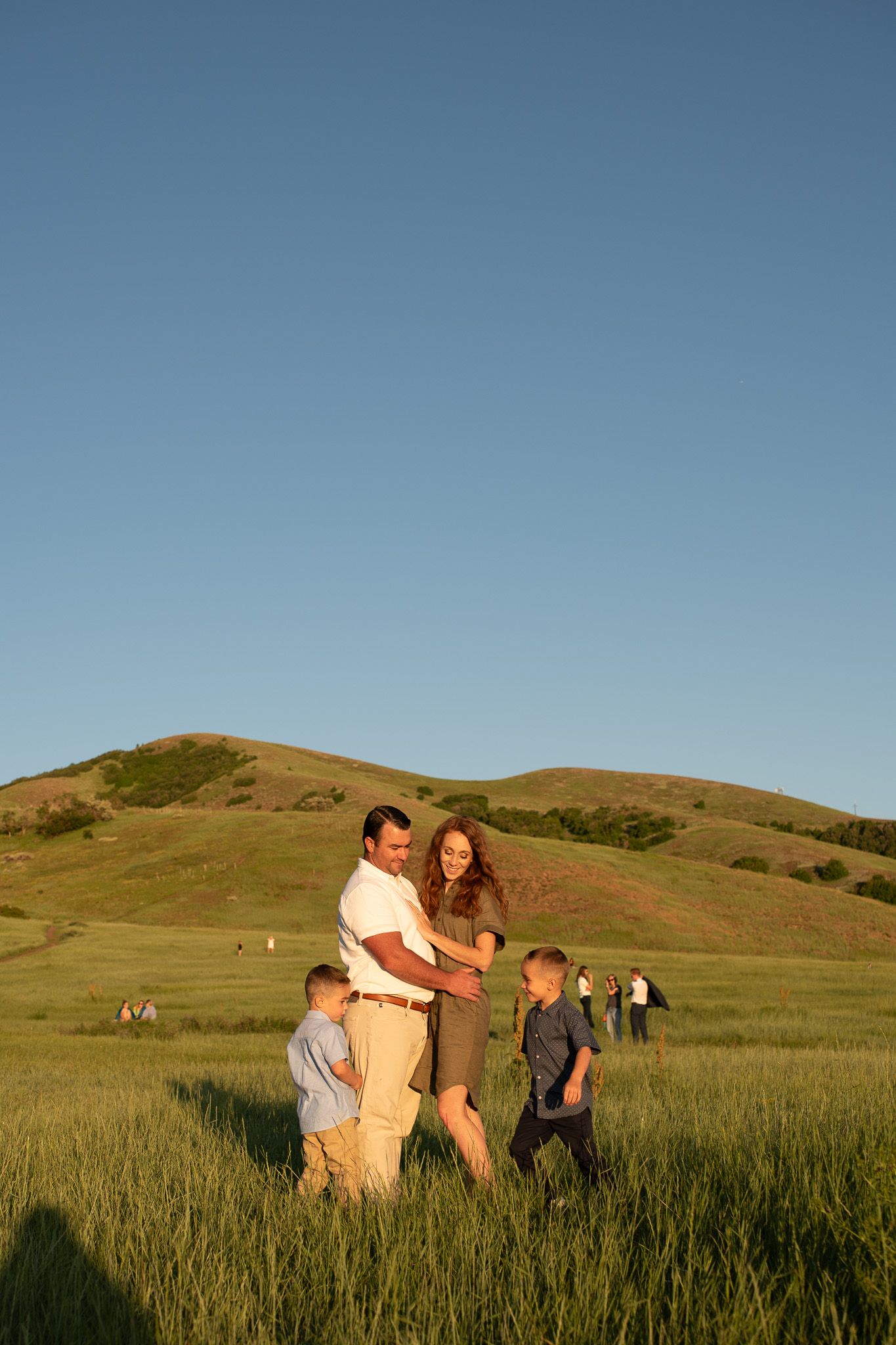 Family of four enjoying golden hour in a green meadow with rolling hills and clear blue sky backdrop.