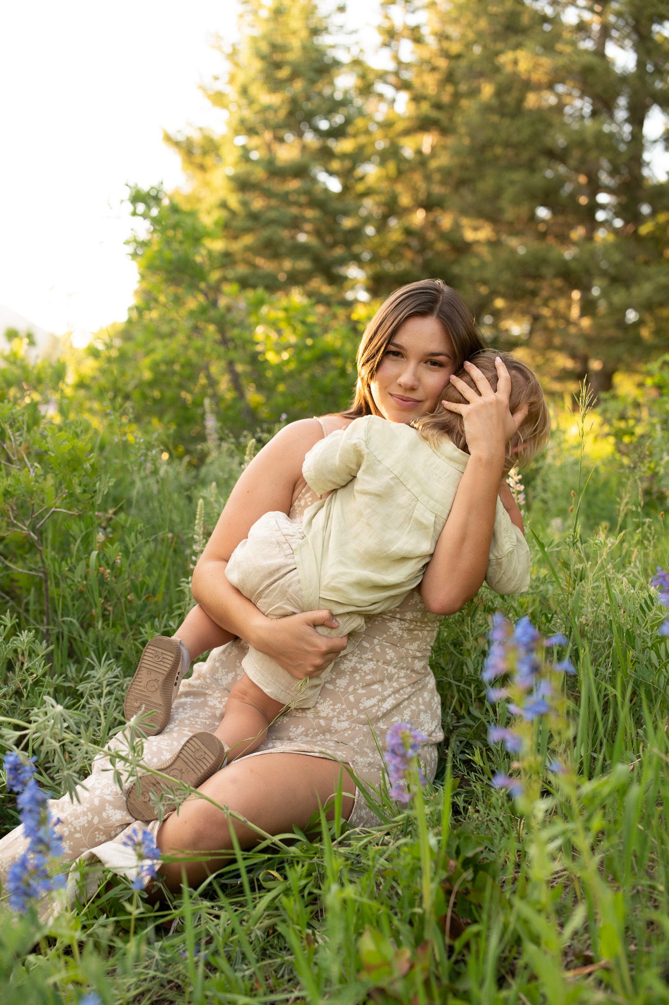 A mother sits in a wildflower meadow cradling her young child, surrounded by green grass and blue lupine flowers.