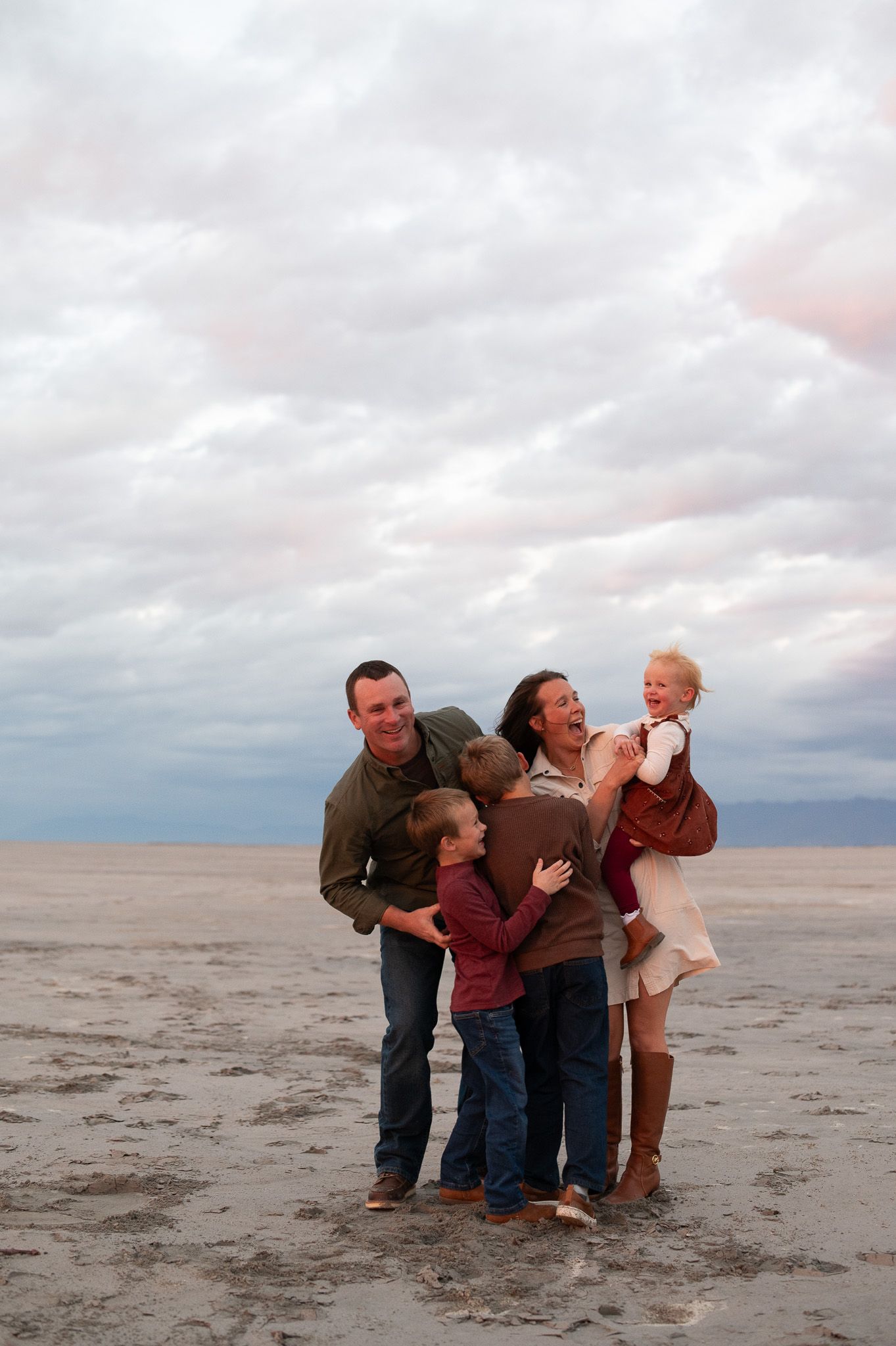 Happy family of four posing on a beach under cloudy skies, parents holding children in a joyful embrace.