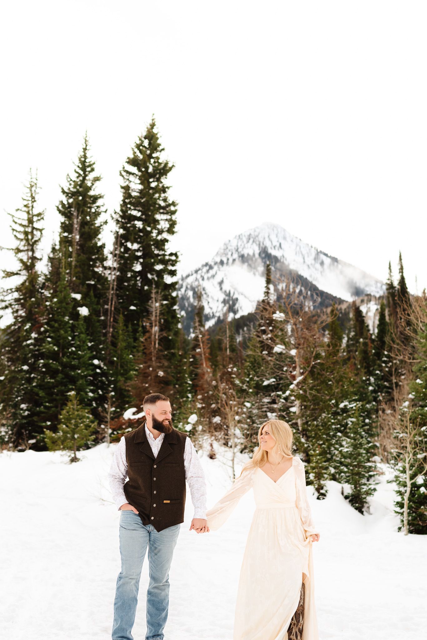 Engaged couple walking through snowy landscape with evergreen trees and mountain peaks in background.
