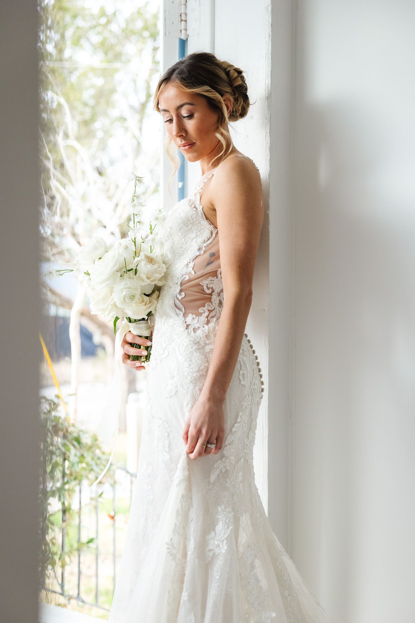 Bride in lace mermaid wedding gown holding white floral bouquet, standing by bright window in elegant bridal portrait.