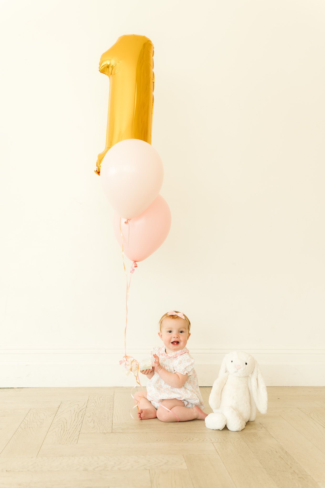 Baby girl celebrates first birthday with gold number one balloon, pink balloons, and white bunny stuffed animal.