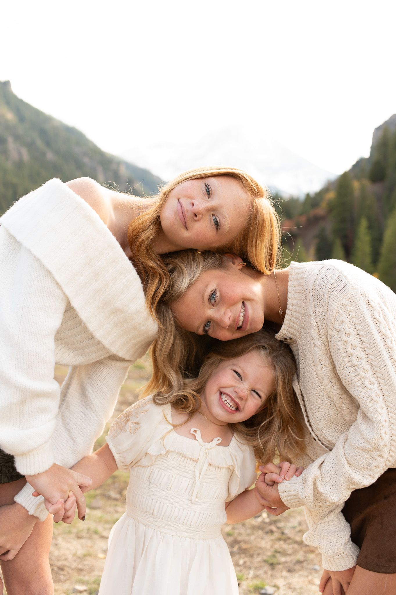 Three blonde females, two women and a young girl, smile and hug outdoors with mountains and trees behind them.