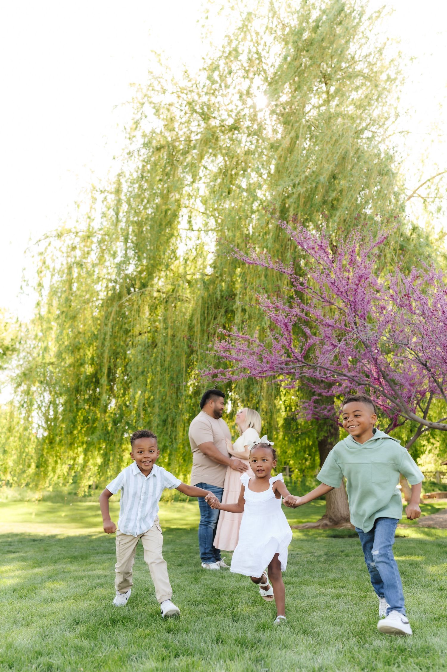 A joyful family plays and runs together on green grass with a weeping willow and flowering tree in the background.